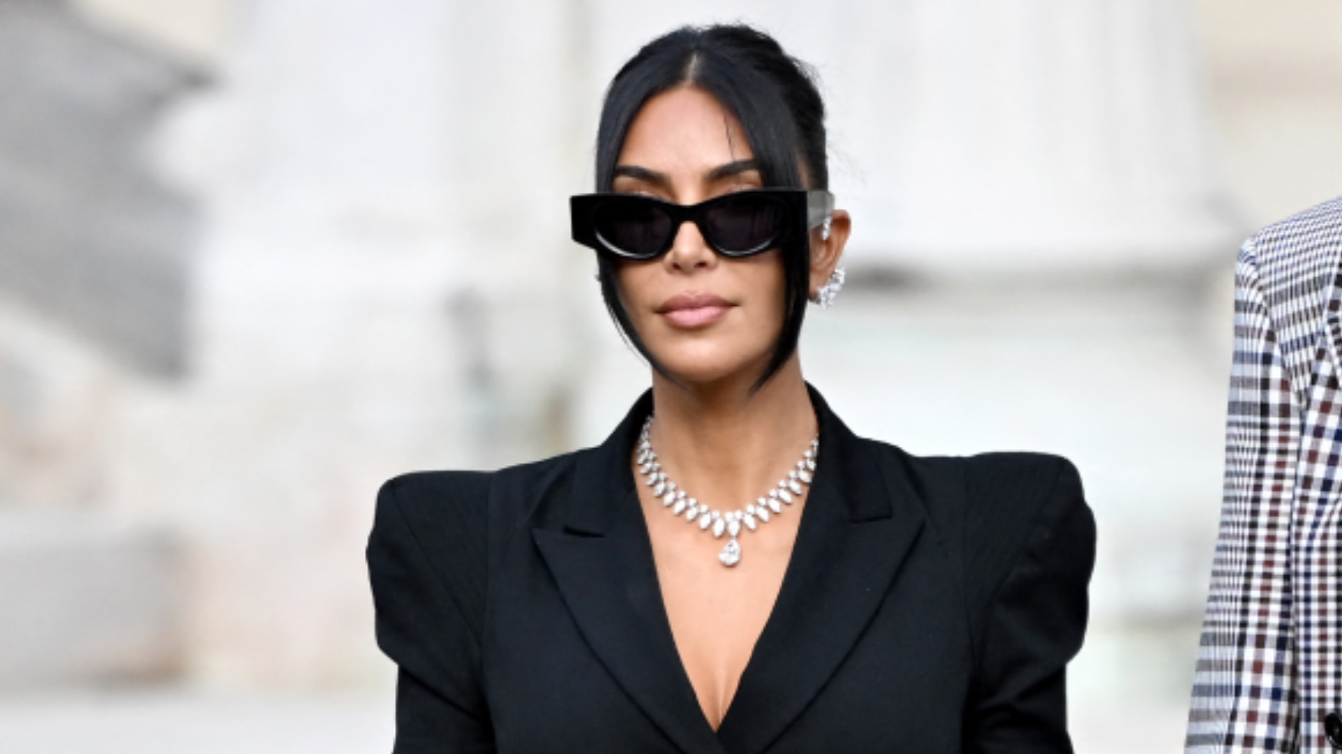 Kim Kardashian wearing sunglasses and a black outfit with a diamond necklace, standing outdoors.