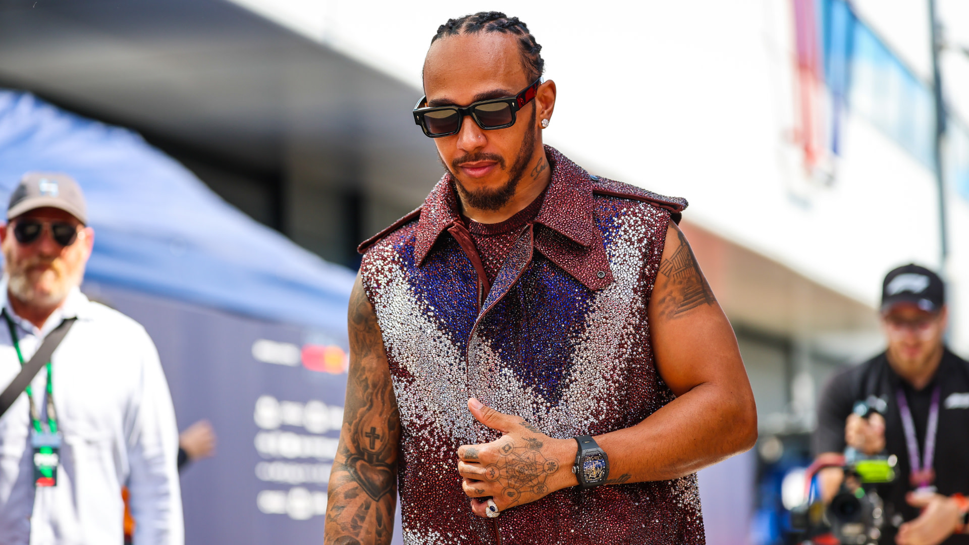 Lewis Hamilton wearing sunglasses and a sparkly sleeveless top, walking at an outdoor event.