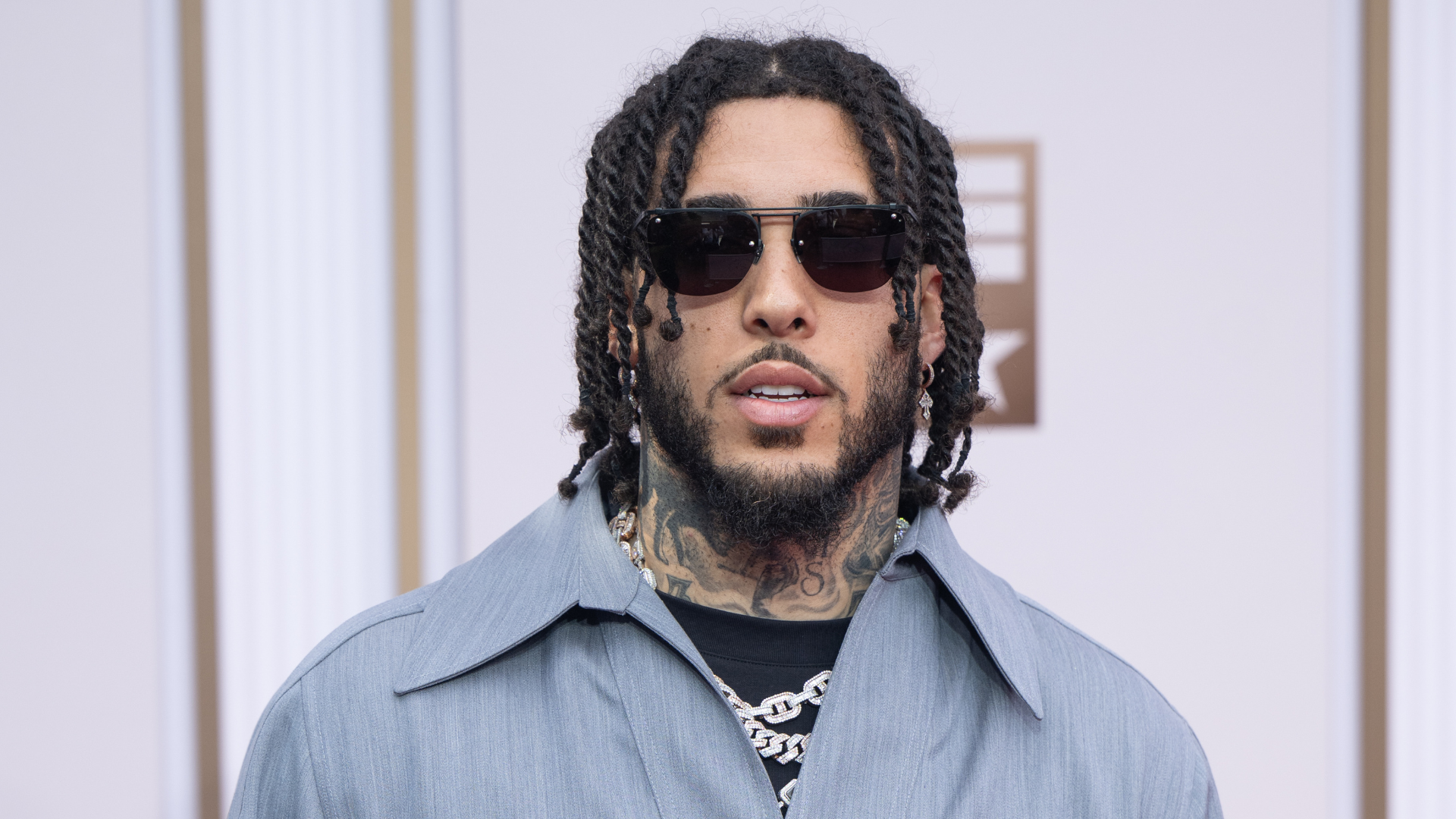 A man with sunglasses, wearing a light blue shirt and chain necklace, stands in front of a patterned background.