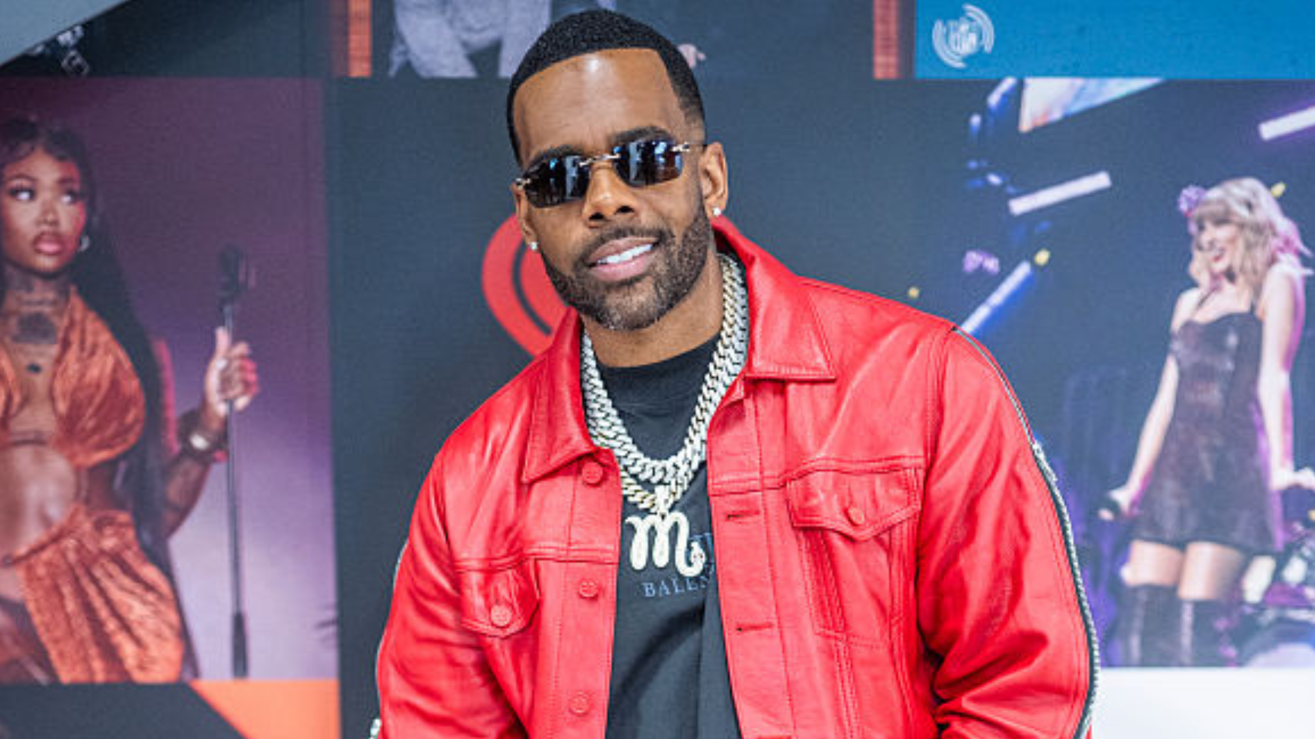 A man in a red jacket and sunglasses poses in front of a backdrop with performers.