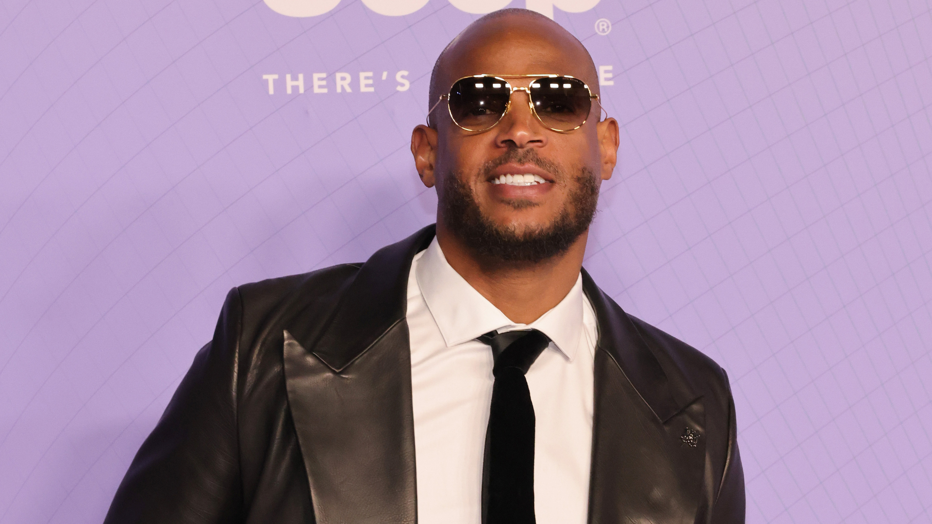 A man wearing sunglasses, a black leather jacket, and a tie, smiling at an event with a purple backdrop.