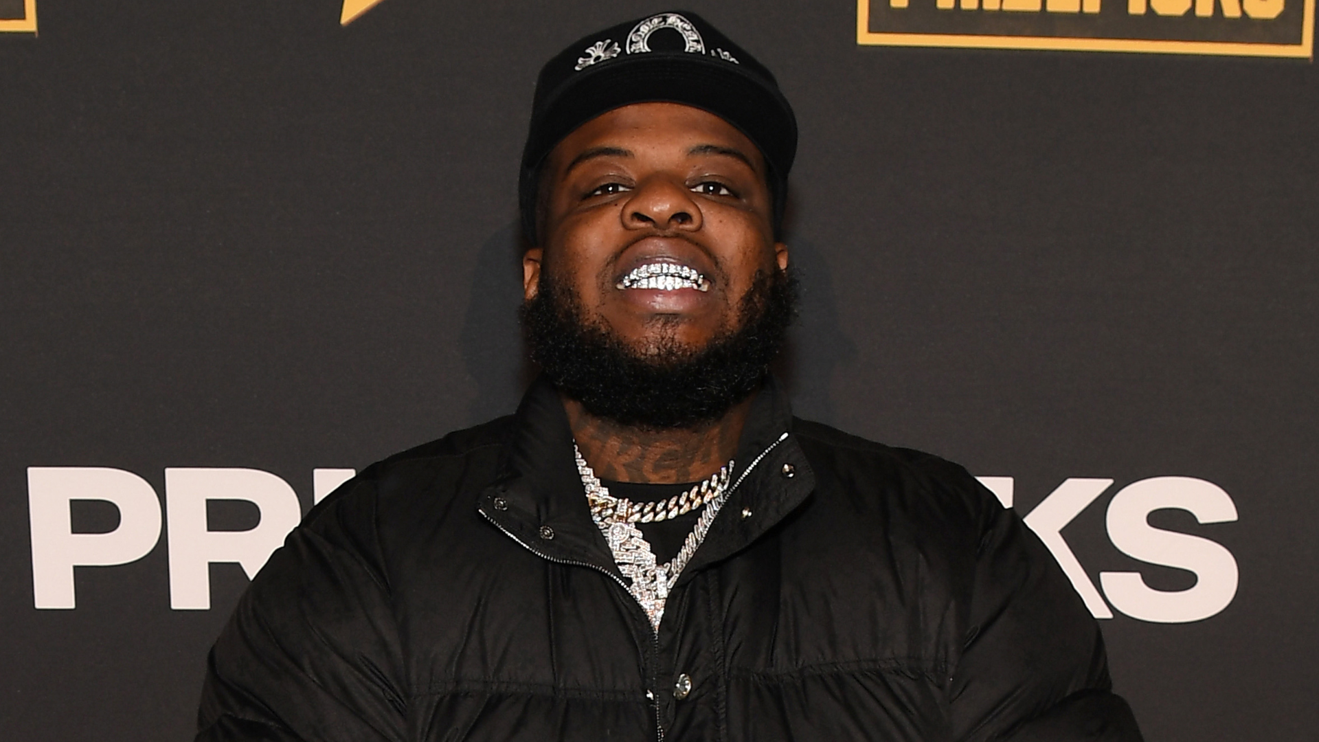 A man with a beard and a cap, wearing a black jacket and silver chains, smiles showing his grillz, against a dark backdrop.