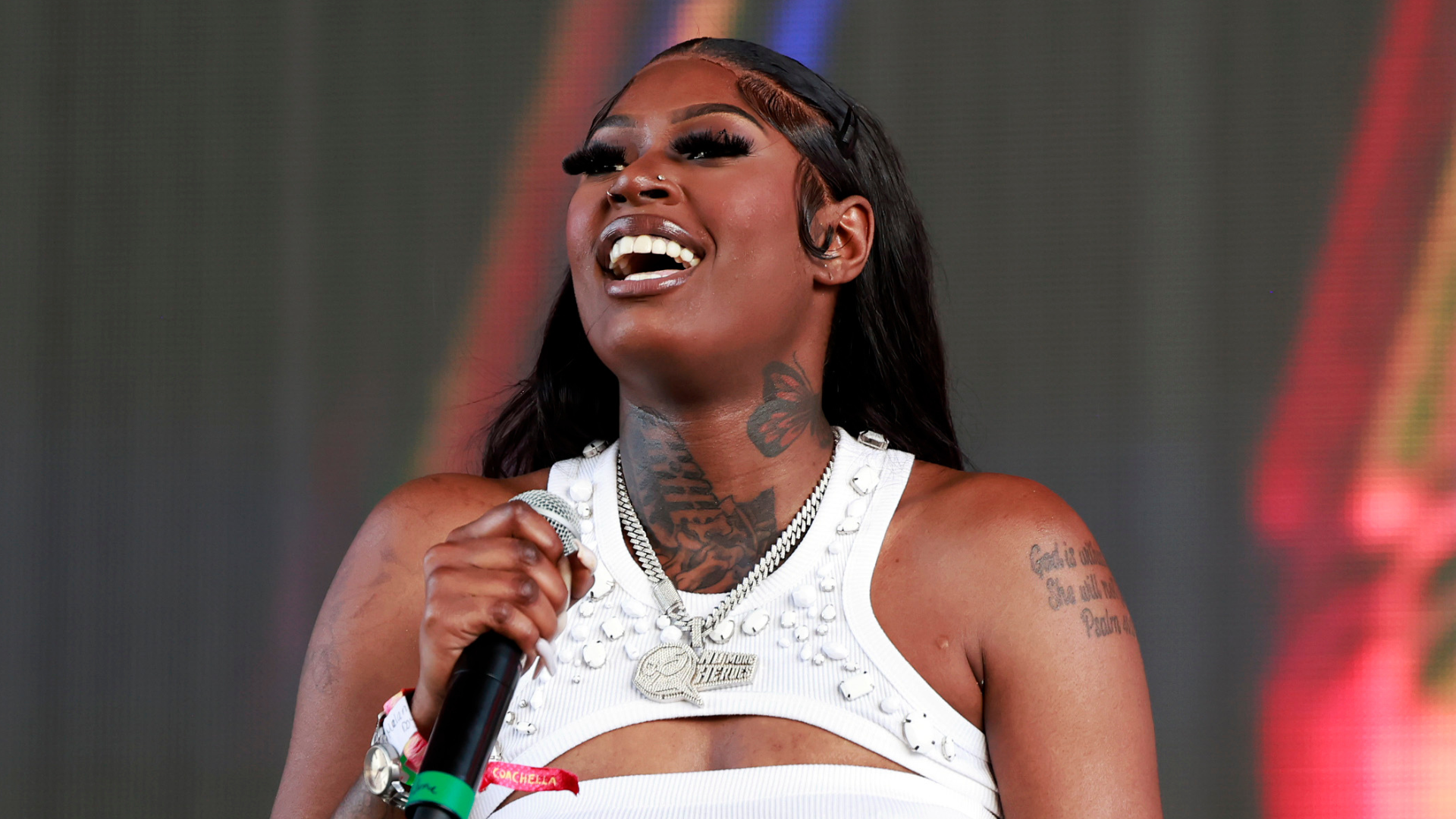Mello Buckzz performing on stage, smiling and holding a microphone. She has long dark hair, a tattoo on her neck, and is wearing a white top.