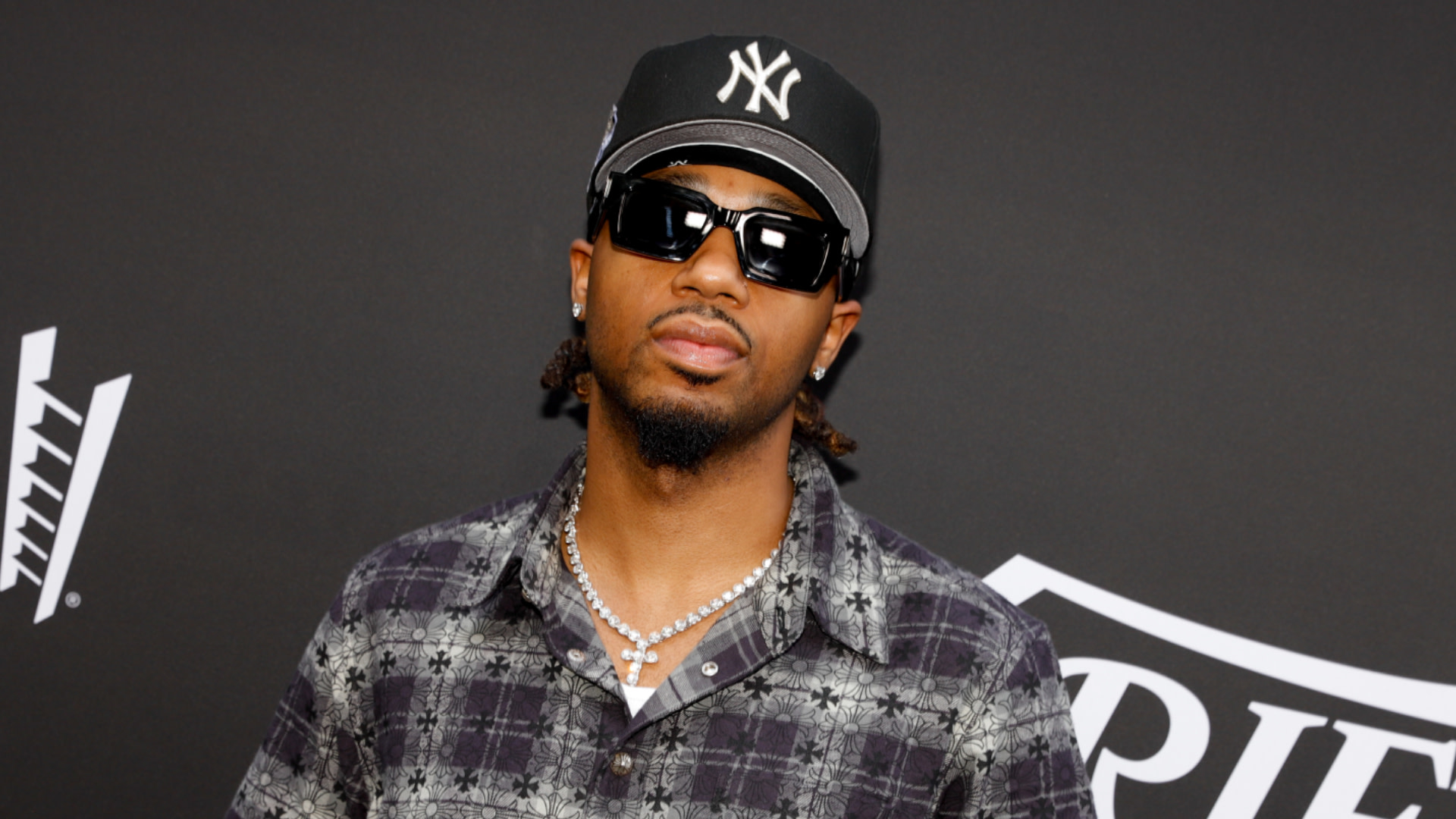Metro Boomin wearing a New York Yankees cap, sunglasses, and a patterned shirt, standing against a dark backdrop.