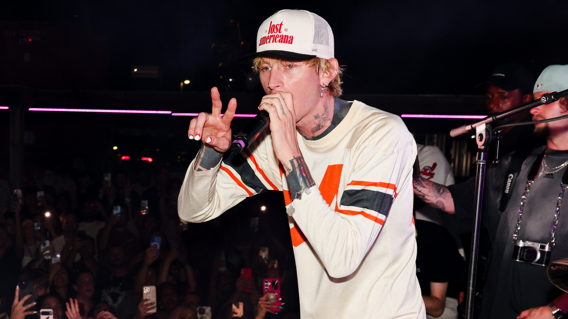 Machine Gun Kelly performing on stage, wearing a white shirt and cap. He's holding a microphone, with a crowd in the background.