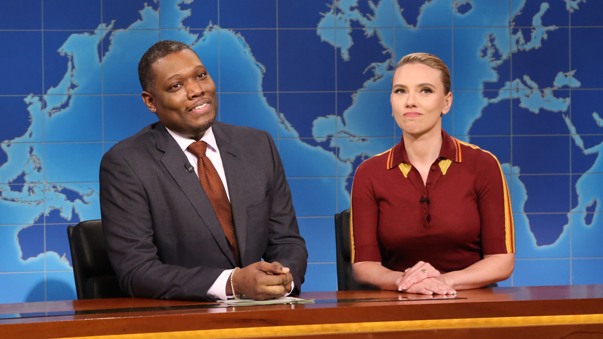 Michael Che and Scarlett Johansson on a TV set with a world map background.