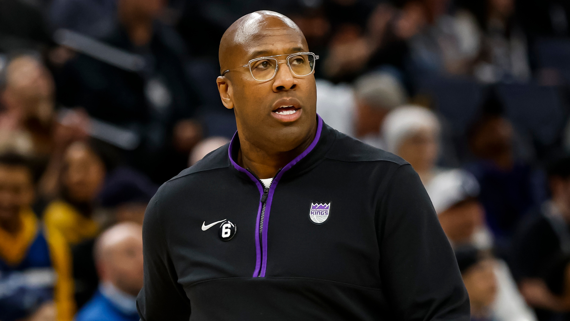 A man wearing glasses and a Sacramento Kings jacket stands in an arena, looking focused.