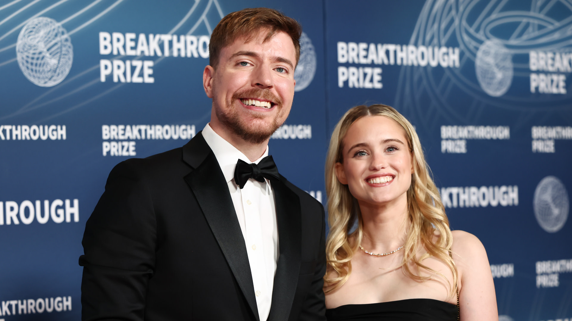 MrBeast in a tuxedo and his fiancée Thea Booysen in a black dress smiling on the red carpet at the Breakthrough Prize event.