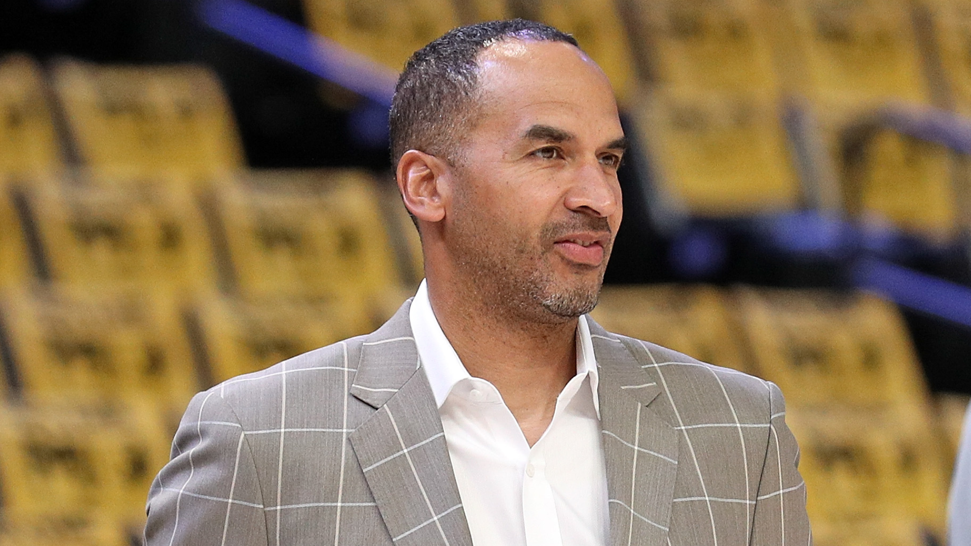 A man in a gray plaid suit stands in an arena with yellow seats.