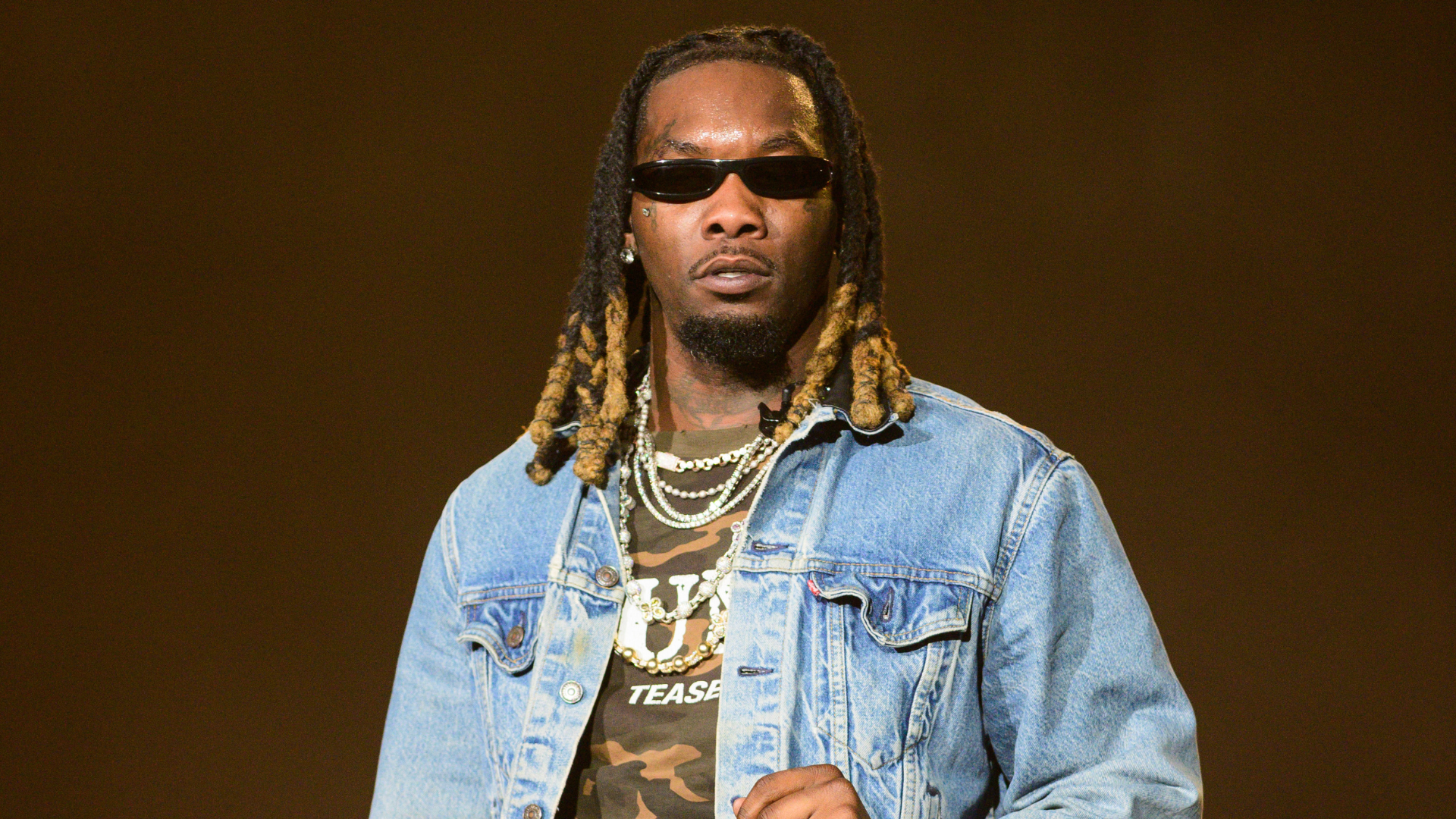 Rapper Offset wearing sunglasses, a denim jacket, and a camo shirt, standing against a plain background.