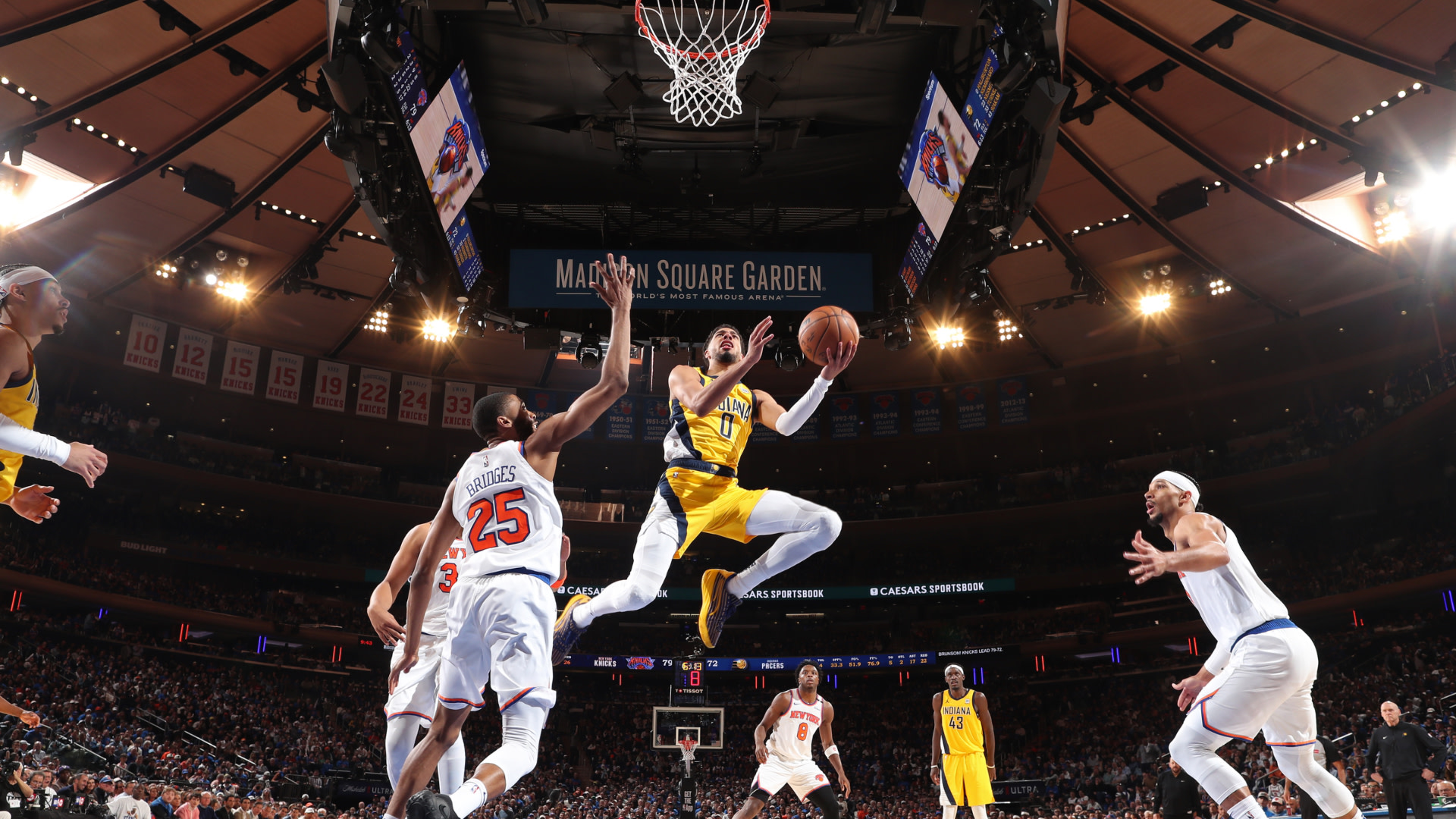 Basketball players in action at Madison Square Garden. A player in a yellow jersey leaps with the ball, surrounded by opponents in white.