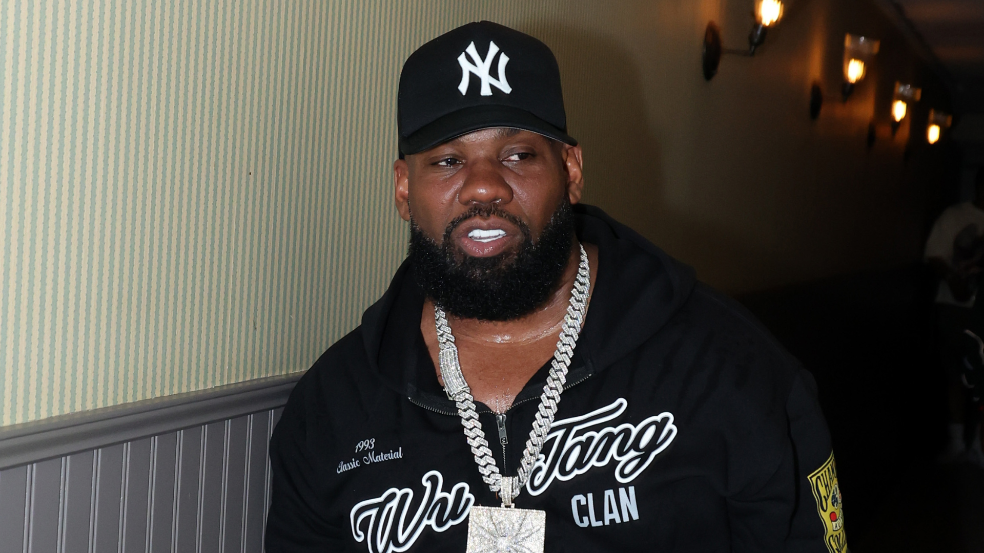 A man wearing a New York Yankees cap, a Wu-Tang Clan hoodie, and a large chain necklace, standing in a hallway with dim lighting.