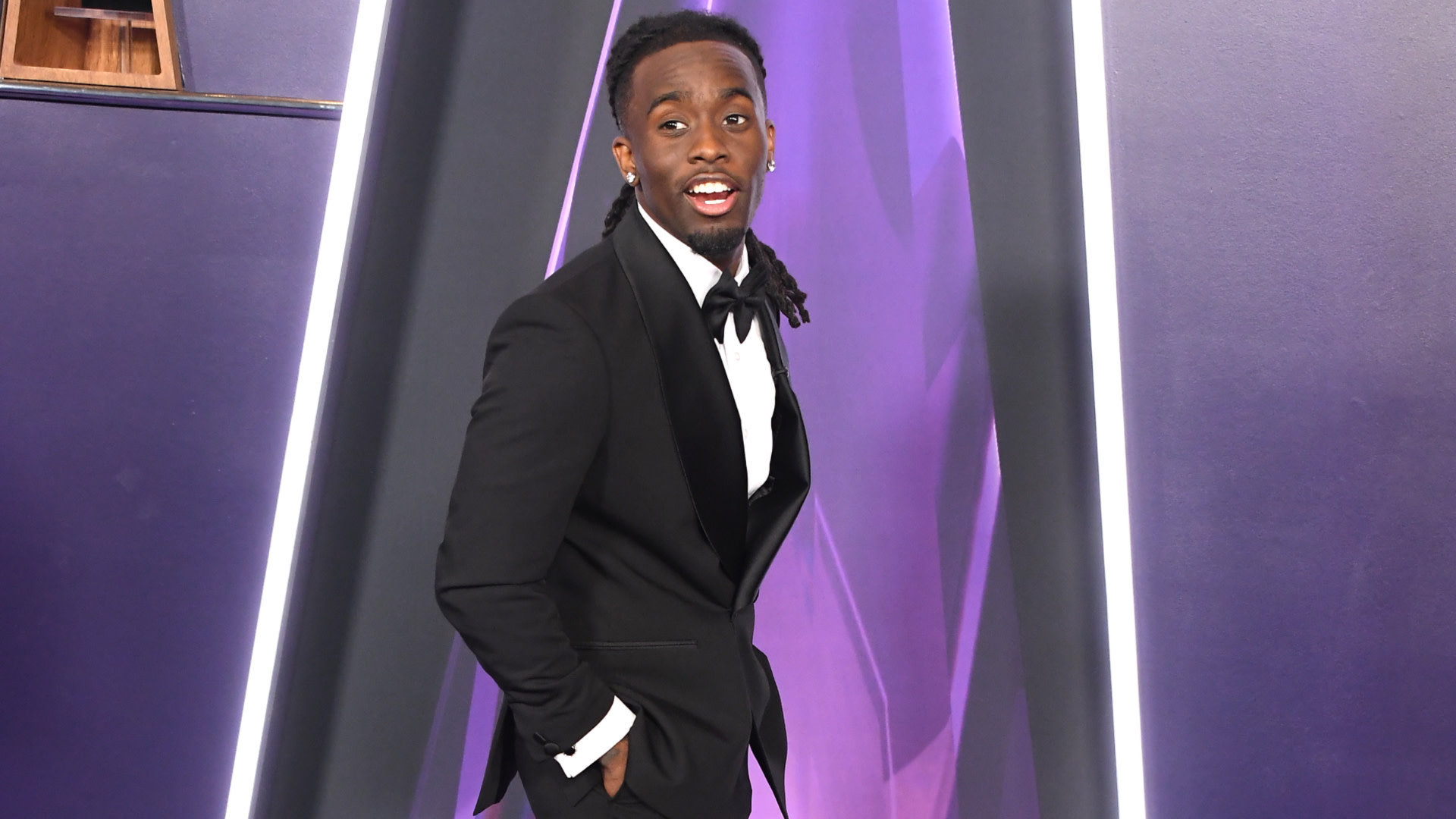 Kai Cenat in a black tuxedo poses on a purple-lit stage, smiling with his hands in his pockets.