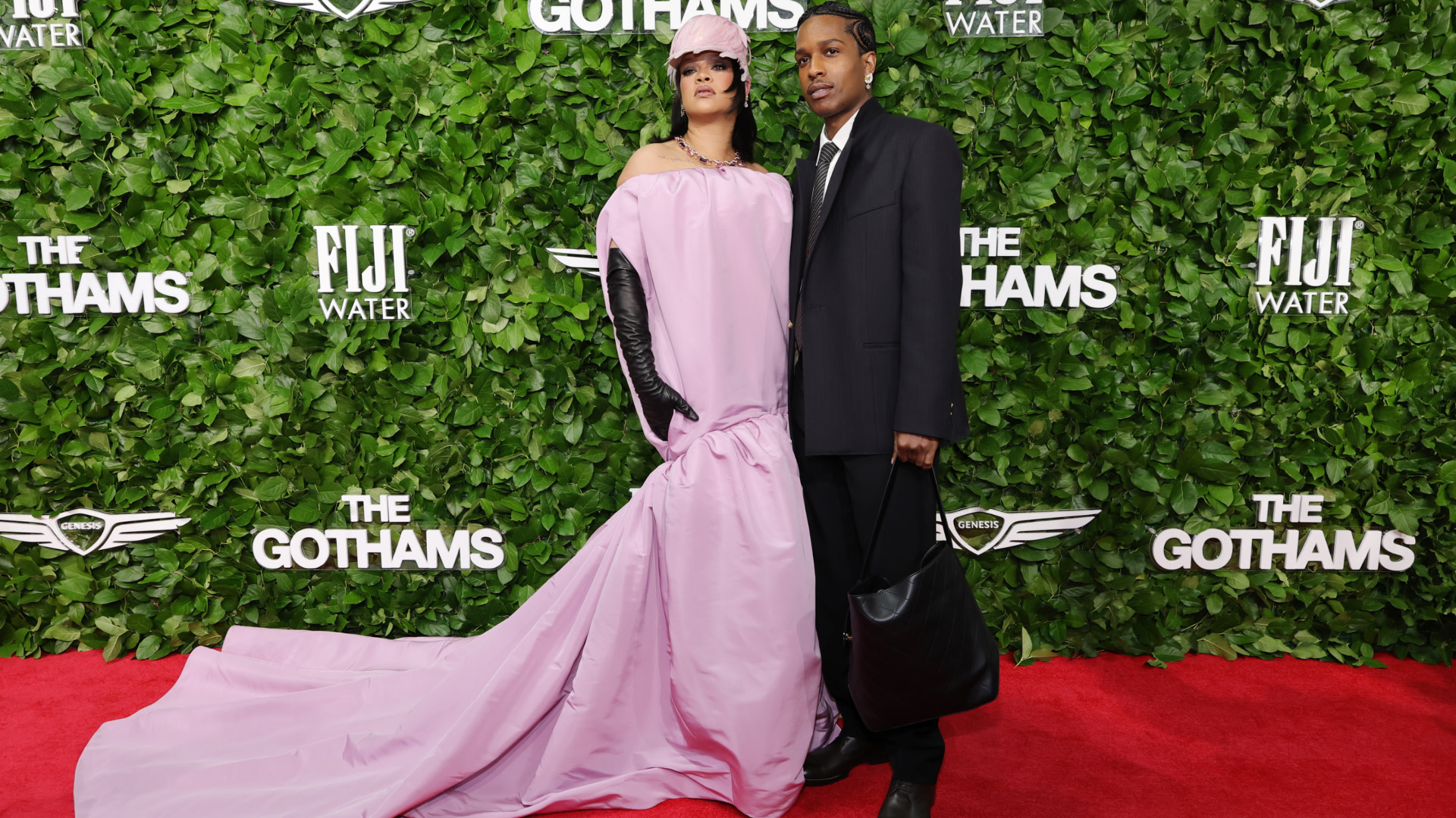 Rihanna in a flowing pink gown and A$AP Rocky in a black suit pose on the red carpet at The Gothams, with a leafy backdrop.