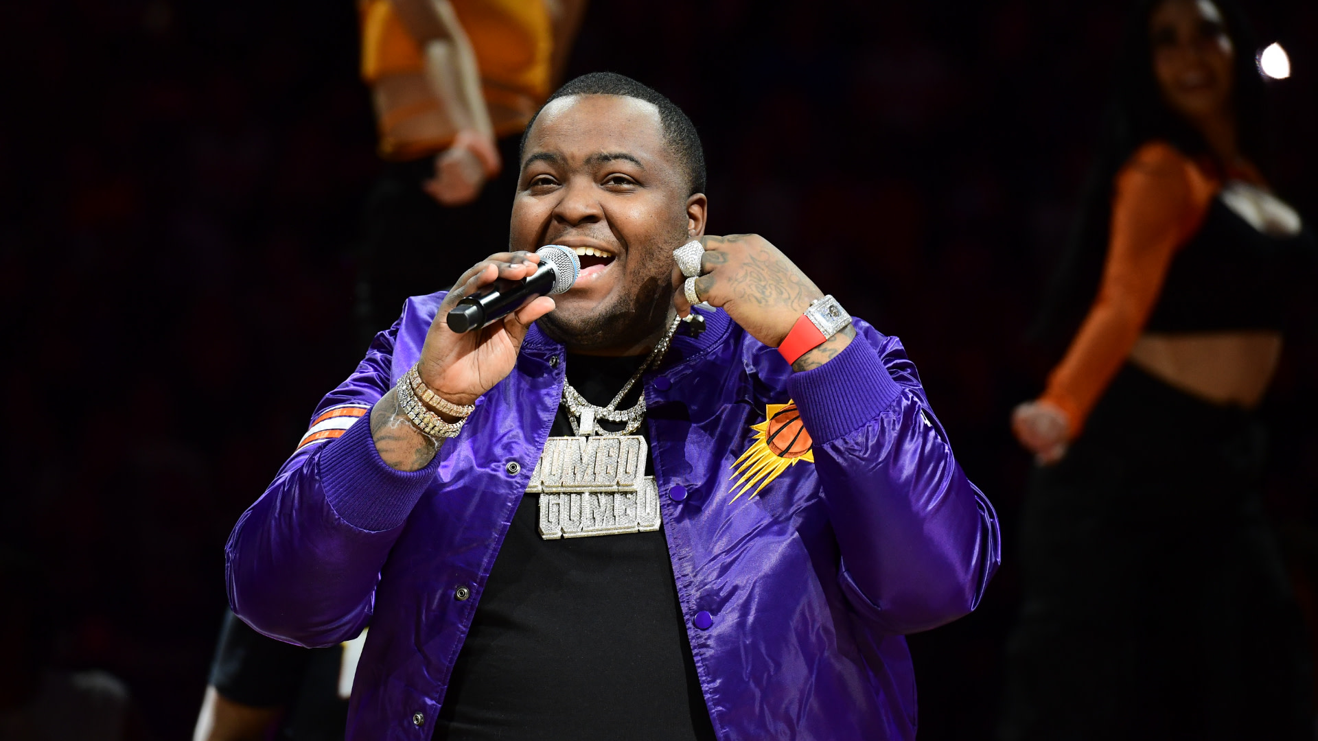 Sean Kingston performs on stage, wearing a purple jacket and holding a microphone, with dancers in the background.