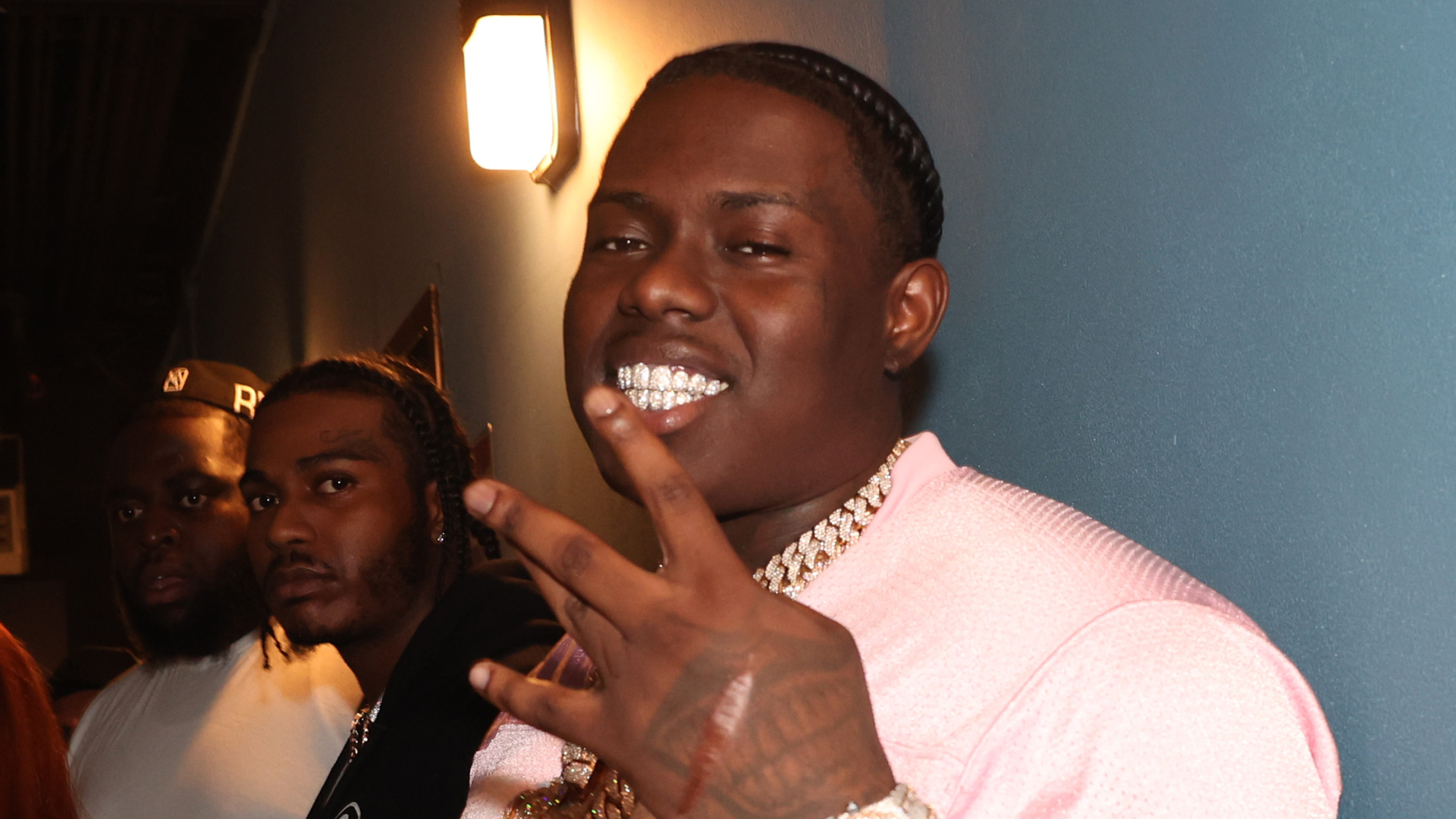 A man in a pink shirt with diamond grills and a chain, smiling and making a hand gesture, with two others in the background.