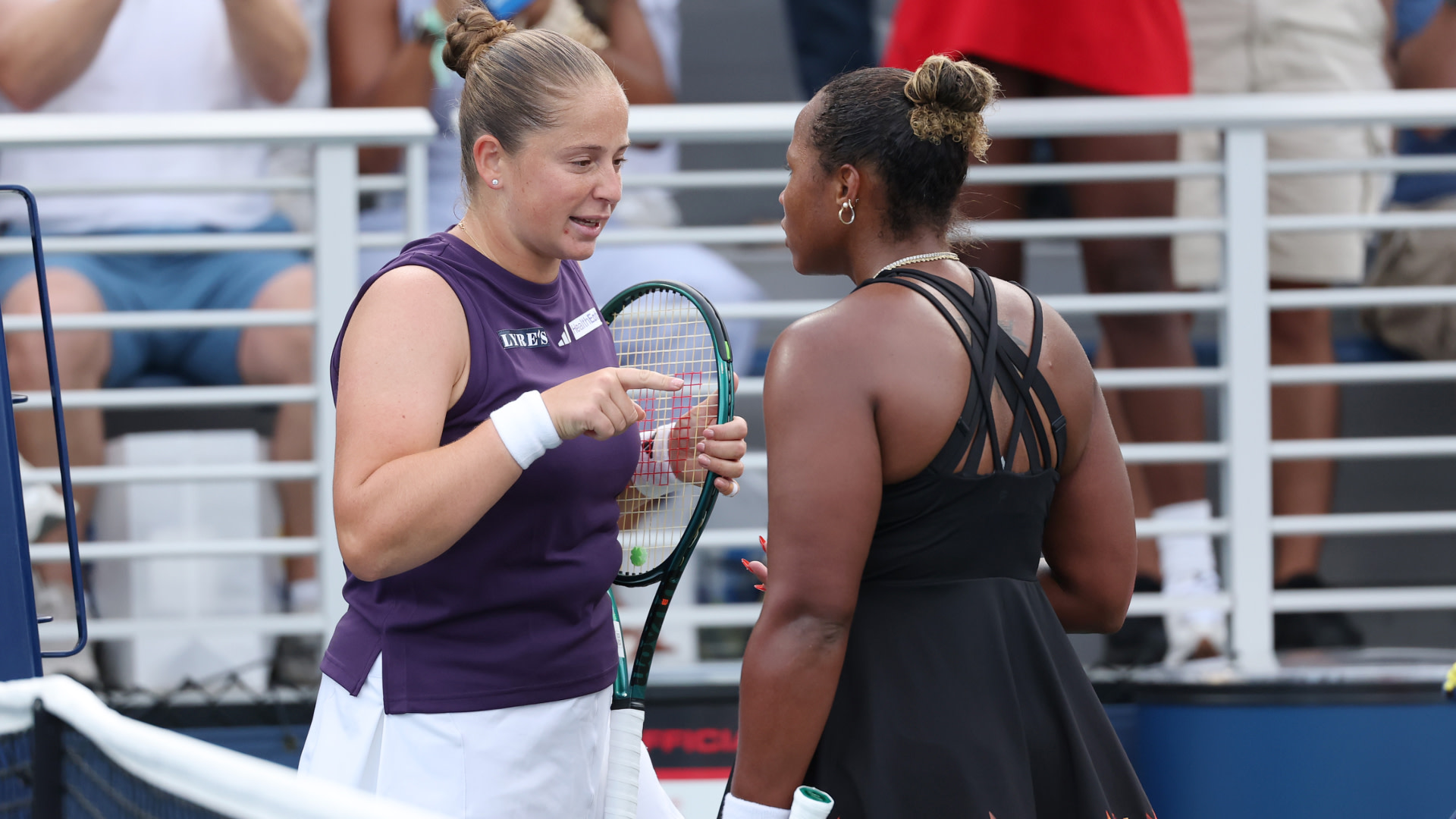 Two female tennis players conversing at the net after a match, with spectators in the background.