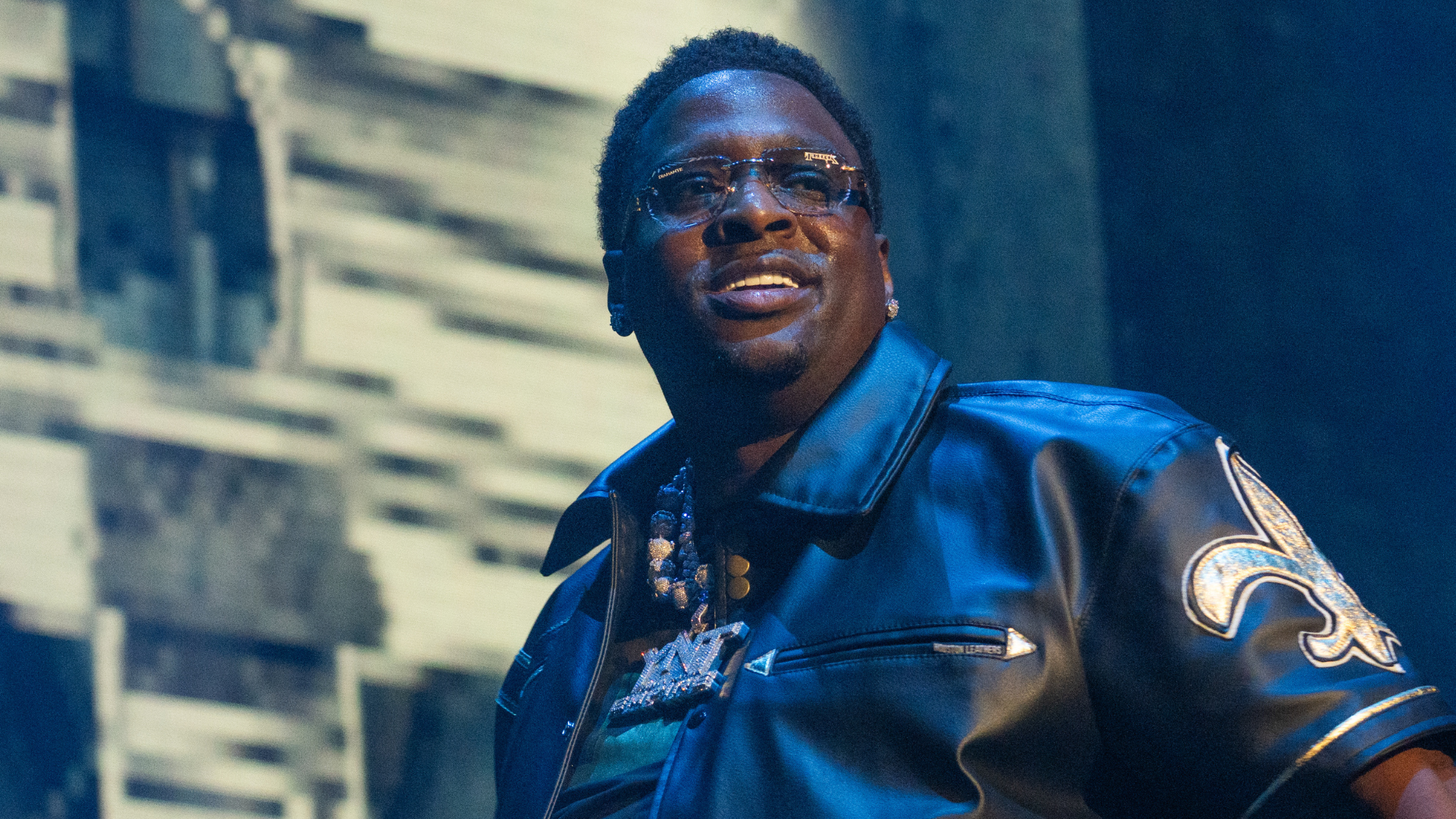 A man wearing glasses and a black leather jacket with a fleur-de-lis emblem, performing on stage.