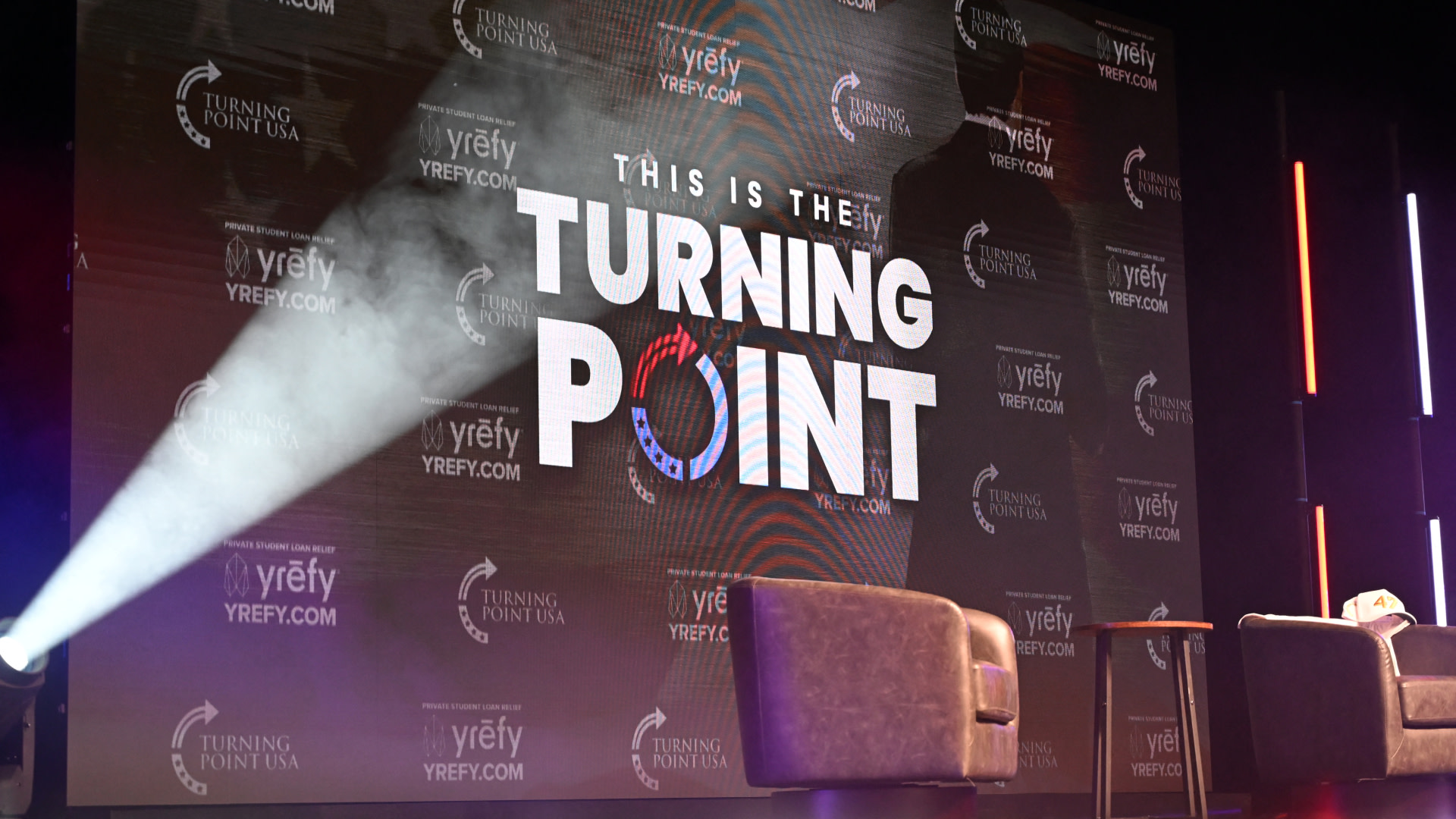 Stage setup for a Turning Point USA event with two chairs, a small table, and a large screen displaying the event's logo.