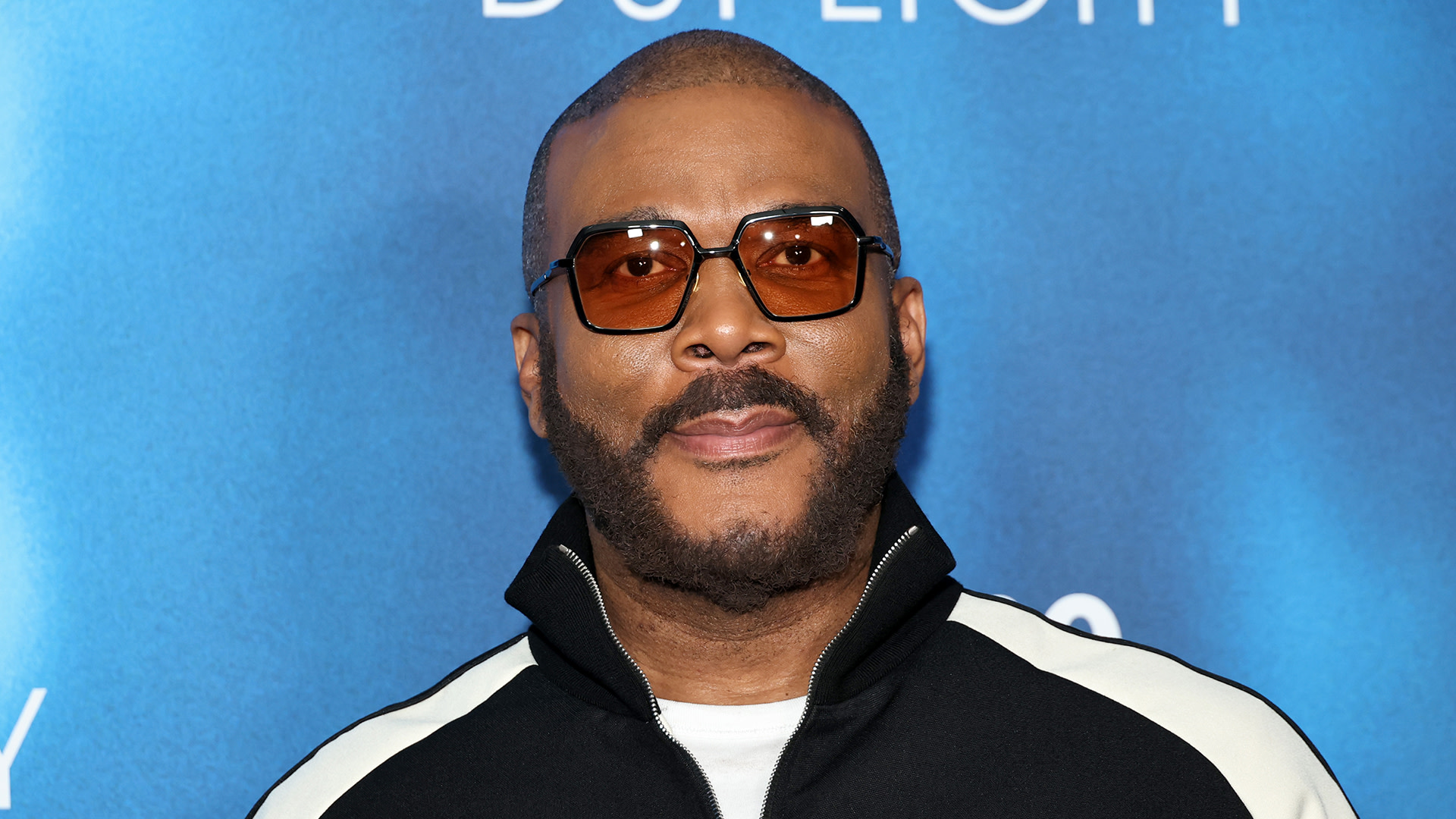 Tyler Perry with glasses and a beard stands against a blue background, wearing a black and white jacket.