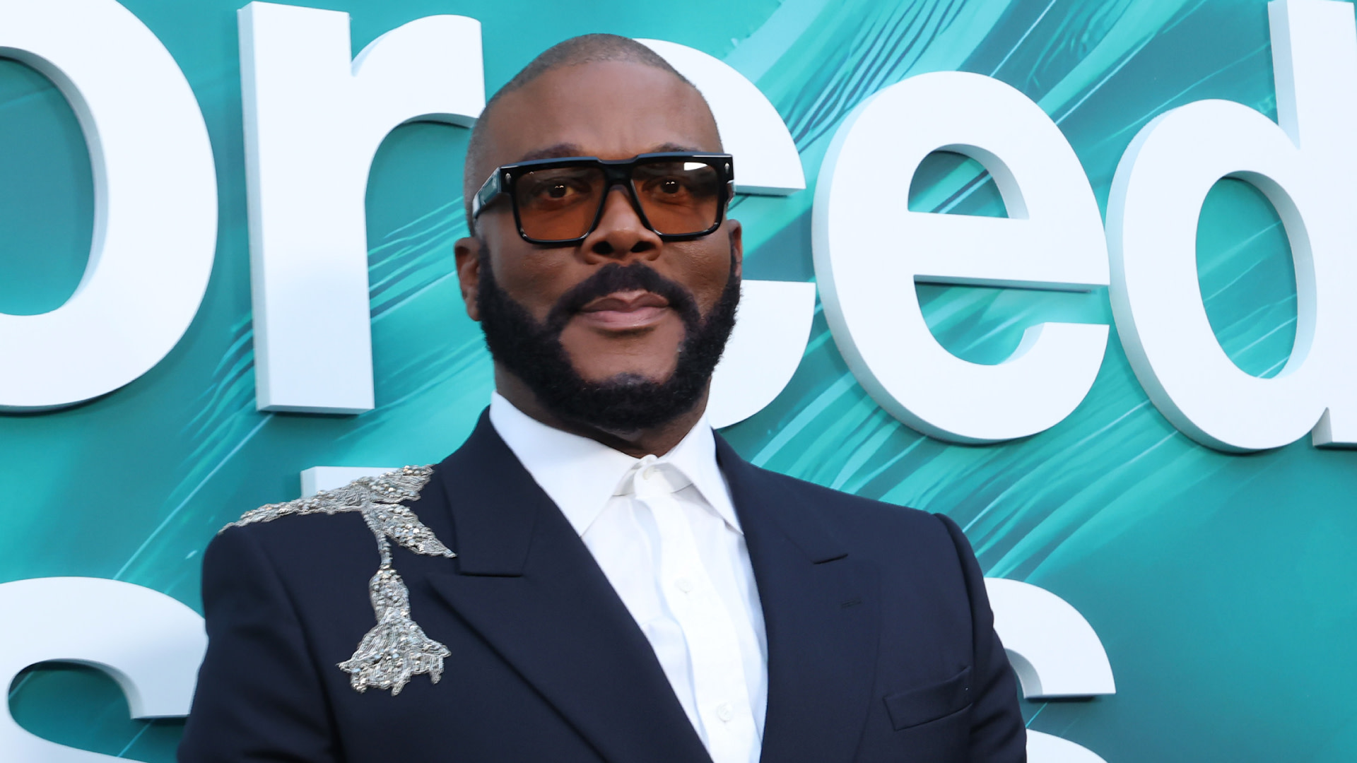 Tyler Perry wearing sunglasses and a dark suit with a decorative brooch, standing in front of a teal backdrop with text.