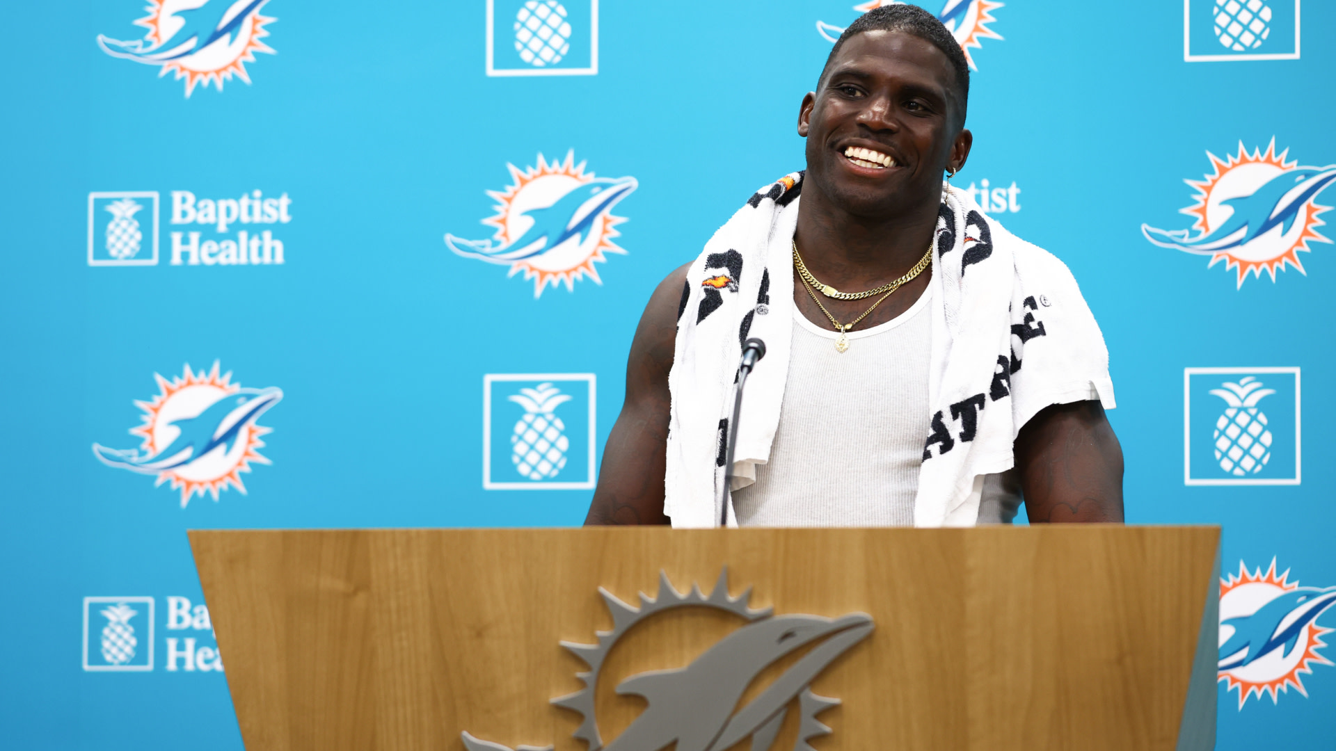 Tyreek Hill stands smiling at a podium with the Miami Dolphins logo, wearing a towel around his neck and a tank top.