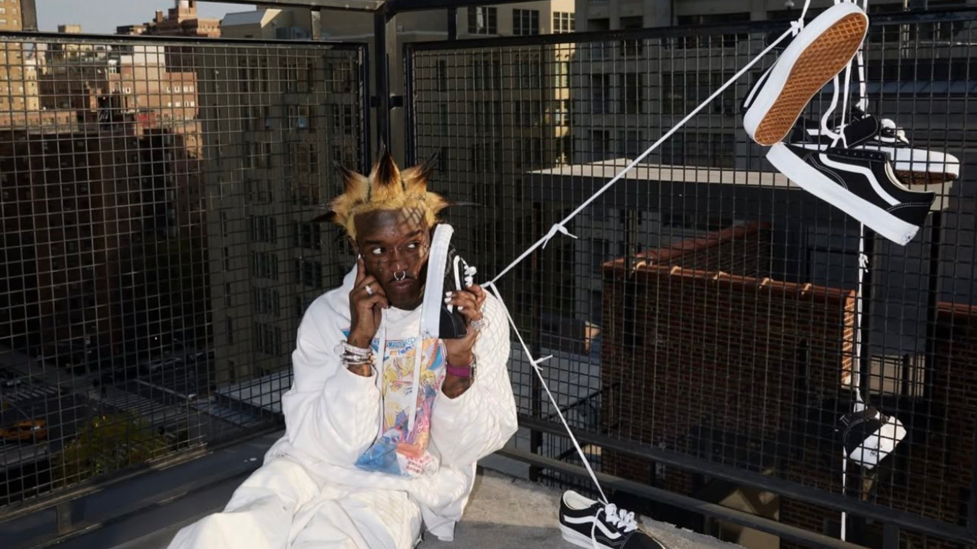 Lil Uzi vert with spiky hair sits on a rooftop, holding a sneaker to their ear like a phone. Several black-and-white sneakers hang nearby.