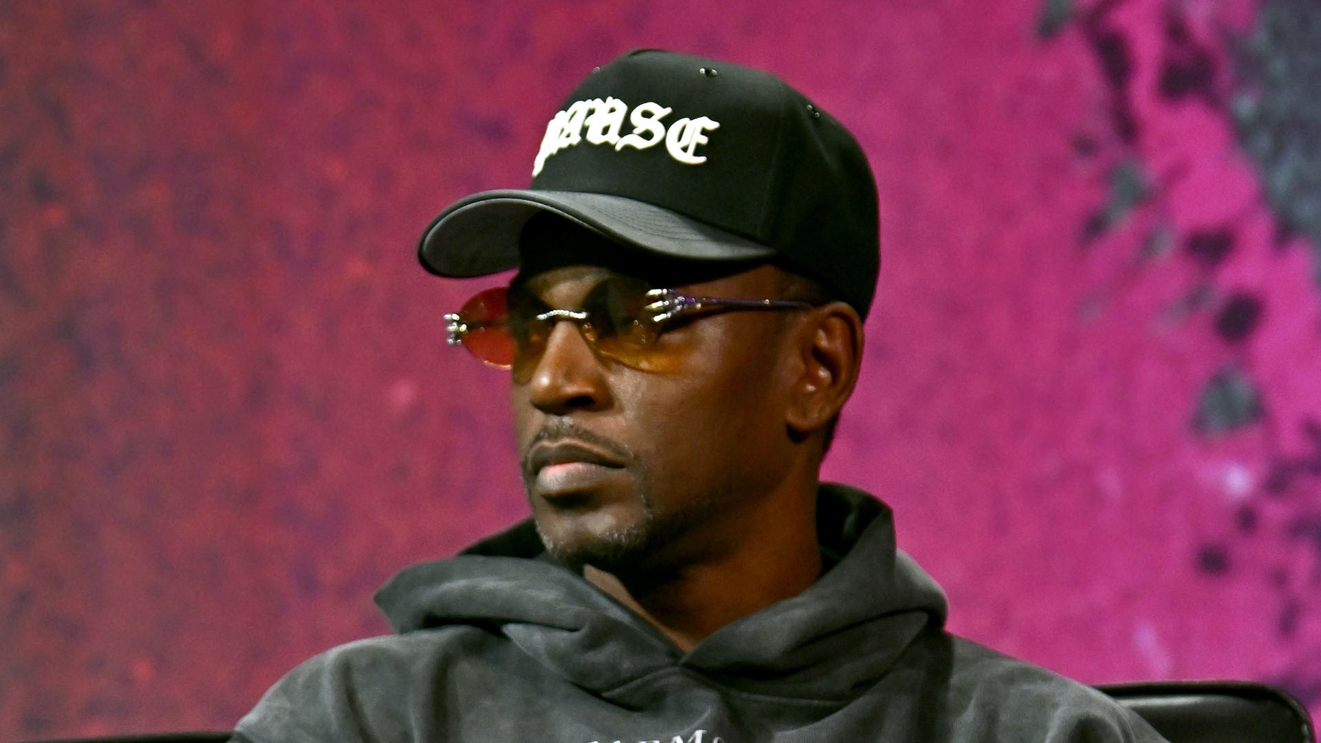 A man wearing a black cap with white lettering, sunglasses, and a gray hoodie sits against a pink background.