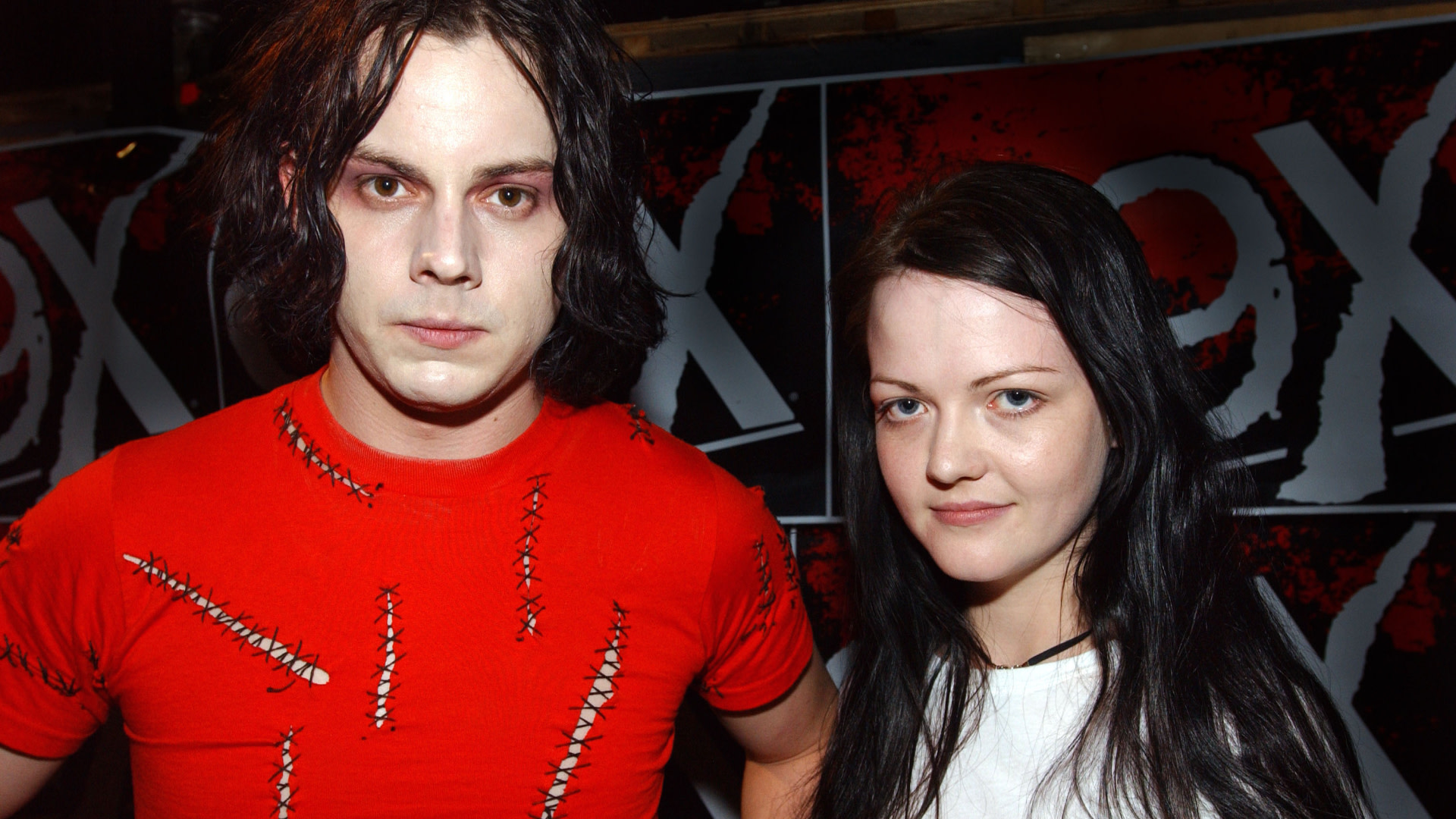 Jack White and Meg White of The White Stripes, standing together. Jack in a red shirt, Meg in white, with a dark background.