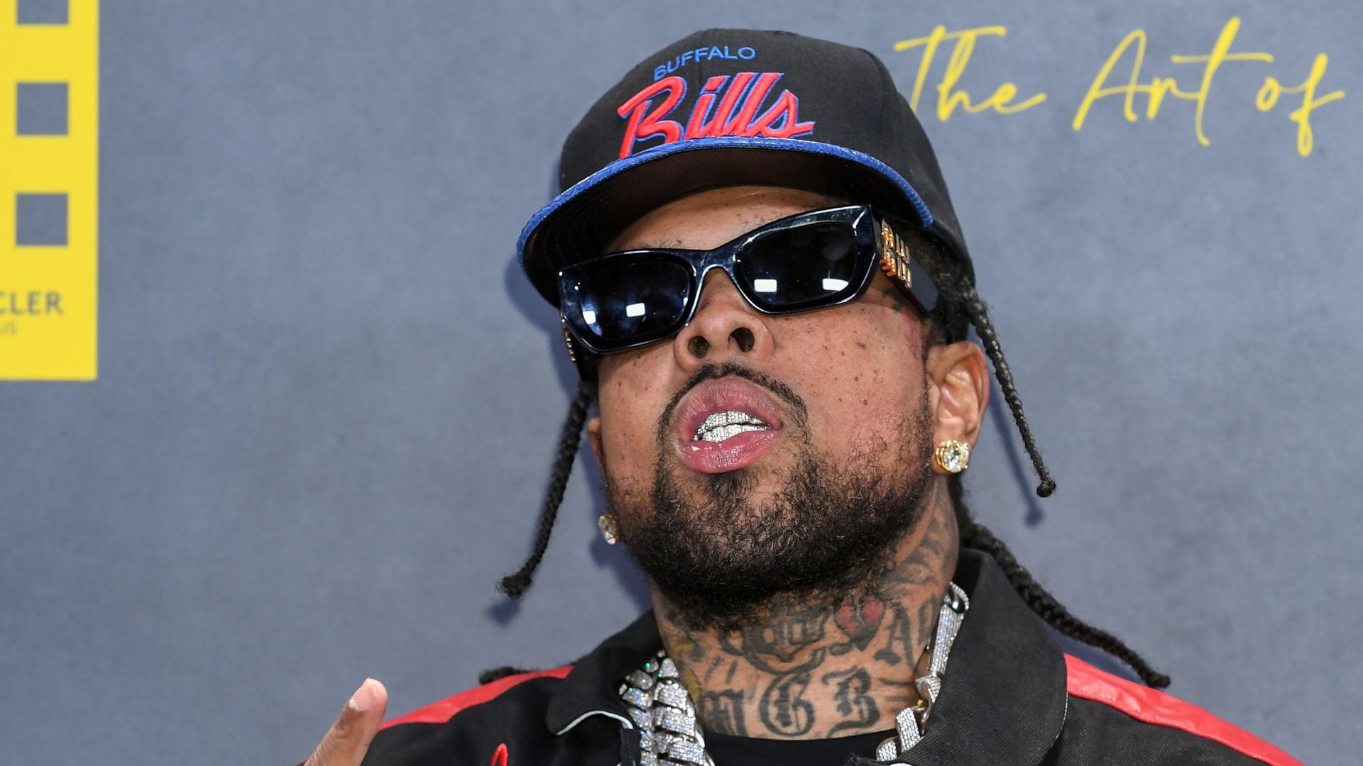 A man wearing a Buffalo Bills cap, sunglasses, and a jacket with a visible tattoo on his neck, standing against a gray background.