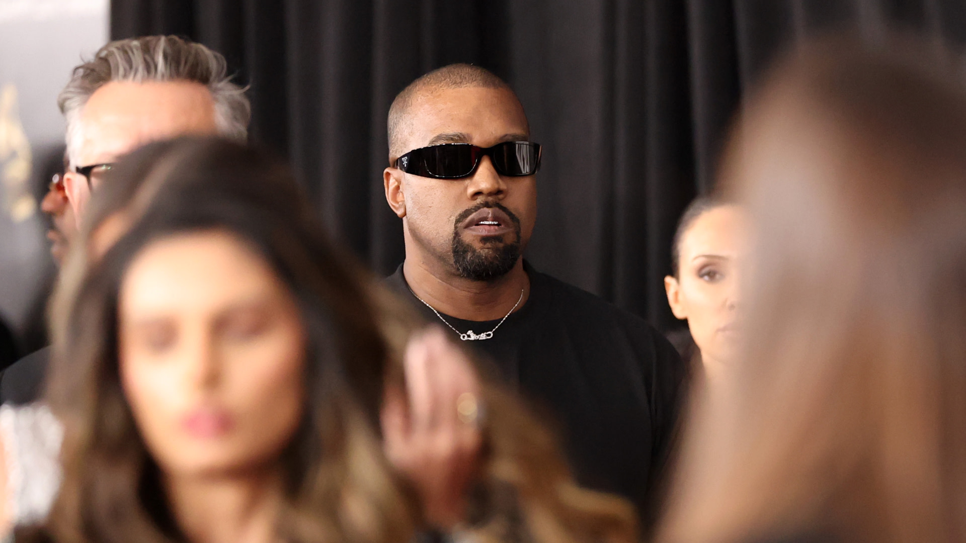 Kanye West wearing sunglasses and a black shirt, stands among a crowd, with a focused expression.