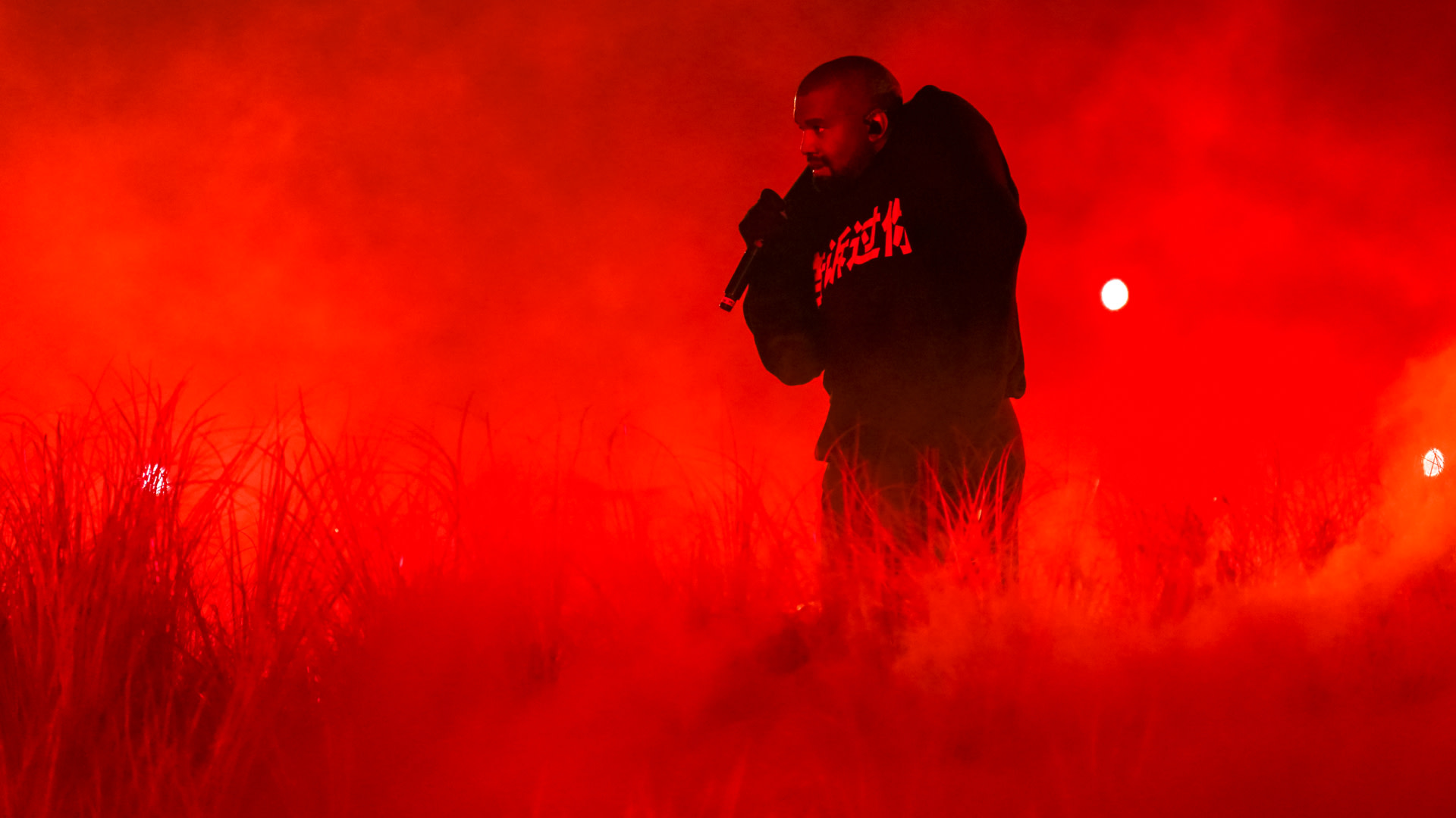 Kanye West performing on stage, surrounded by red lighting and fog, wearing a dark hoodie and holding a microphone.