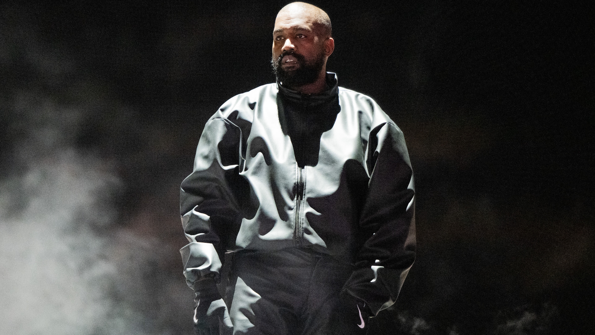 Kanye West standing in a dark setting, wearing a black leather jacket, looking serious.