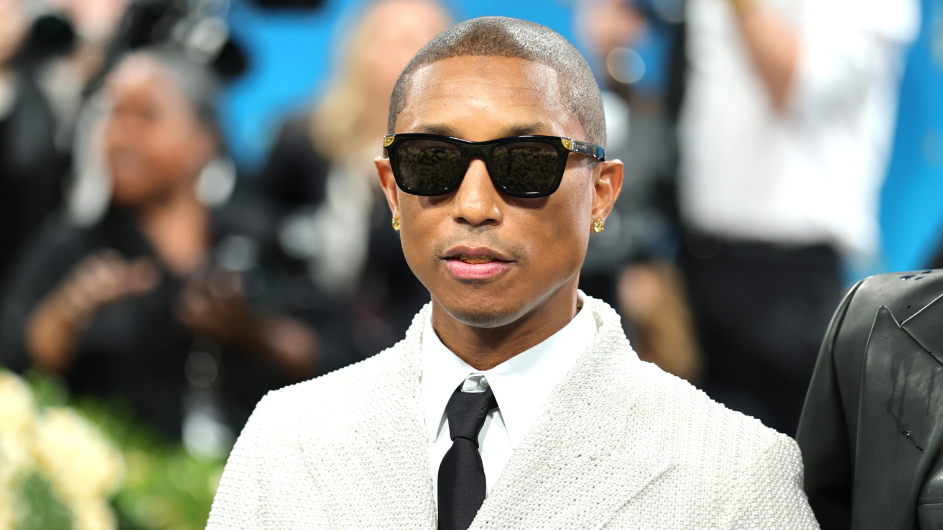 Pharrell Williams in sunglasses and a white textured jacket at a formal event.