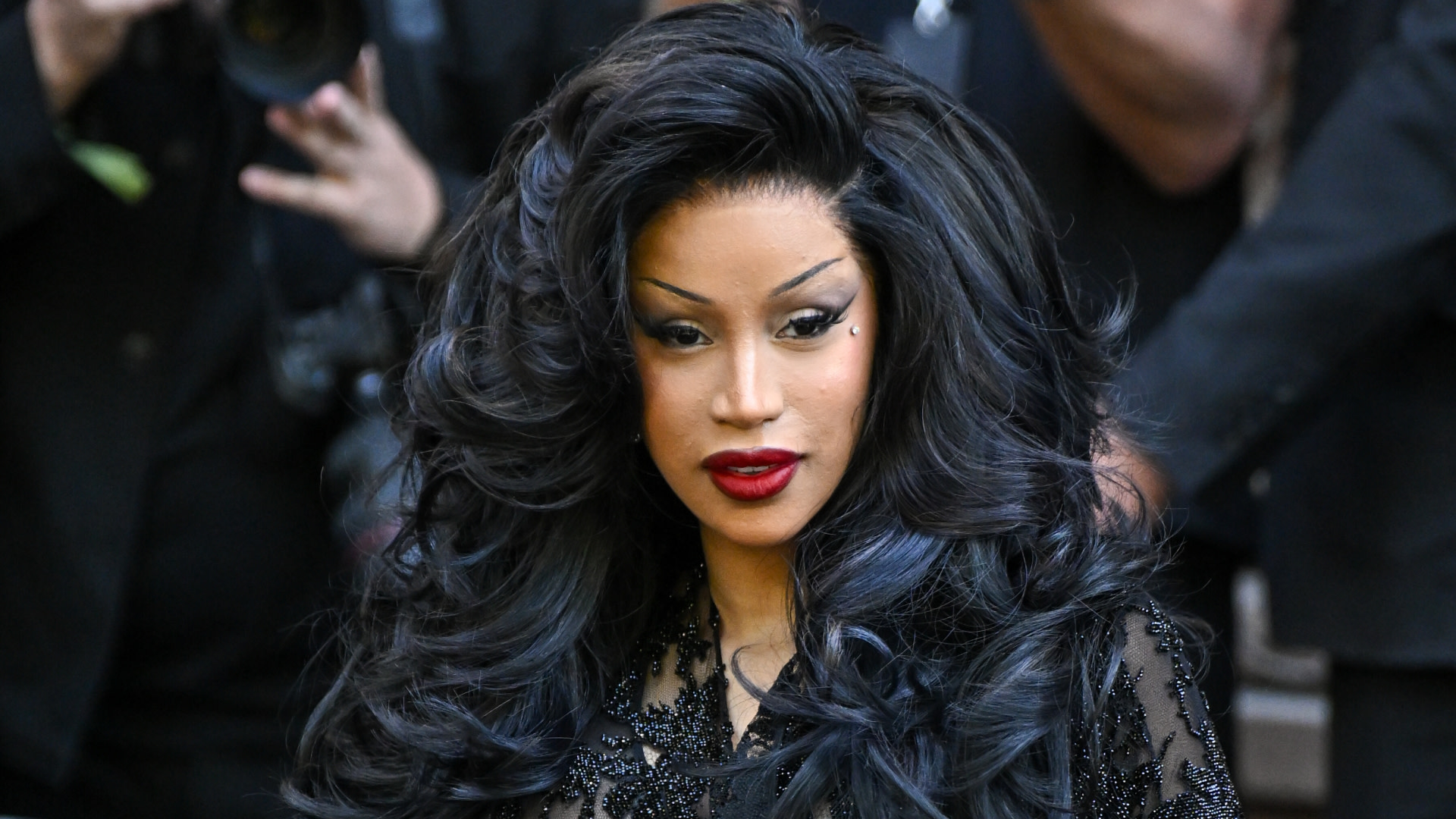 Cardi B with voluminous black hair and red lipstick, wearing a black embellished outfit, in front of a crowd.