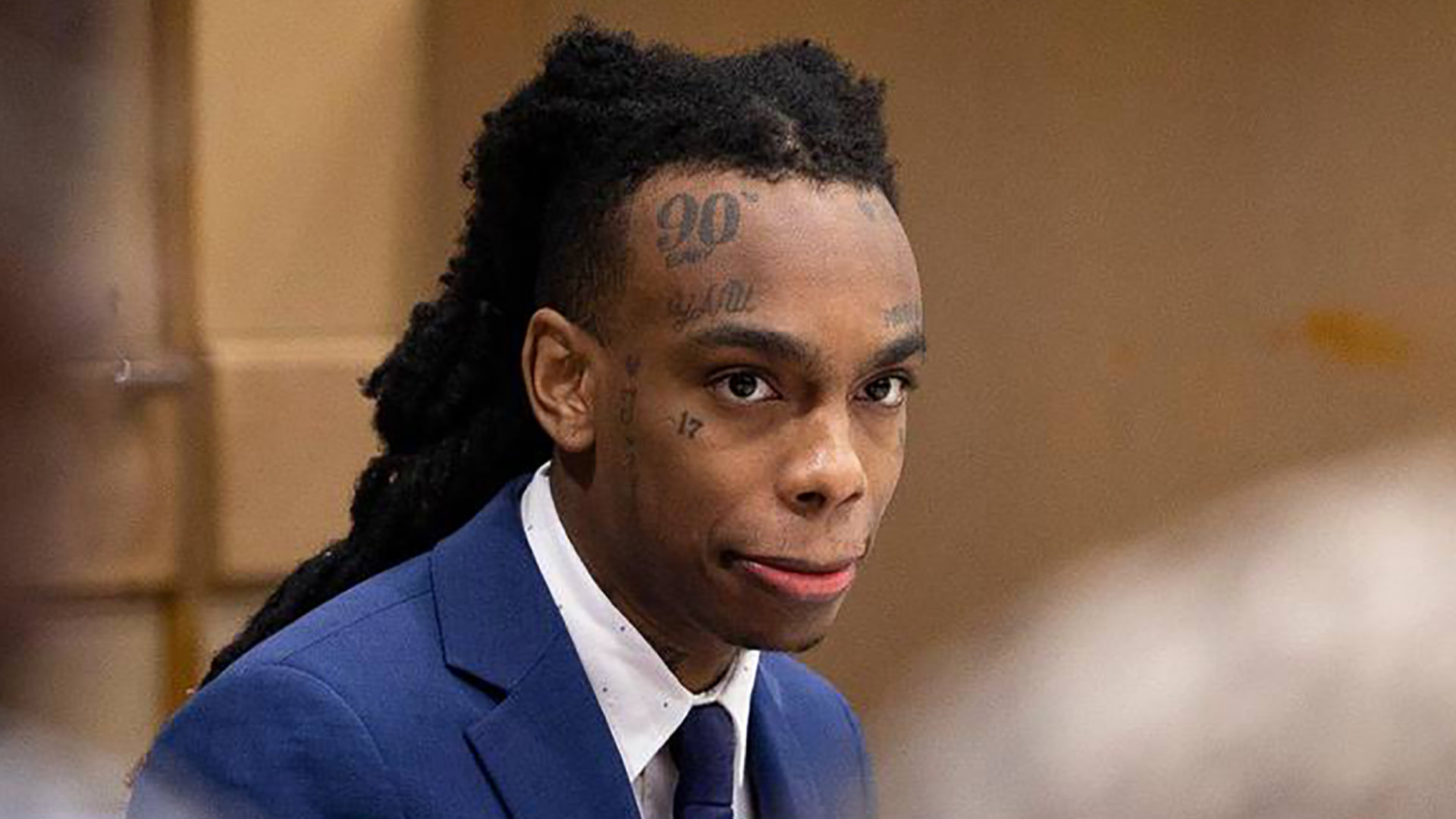 YNW Melly with tattoos on his face, wearing a blue suit and tie, sitting in a courtroom setting.