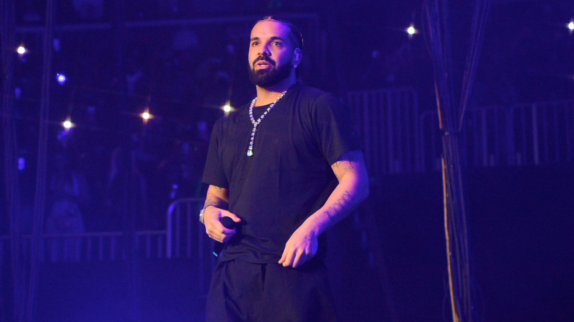 Drake performing on stage, wearing a black shirt and a necklace, with a spotlight on him and a crowd in the background.