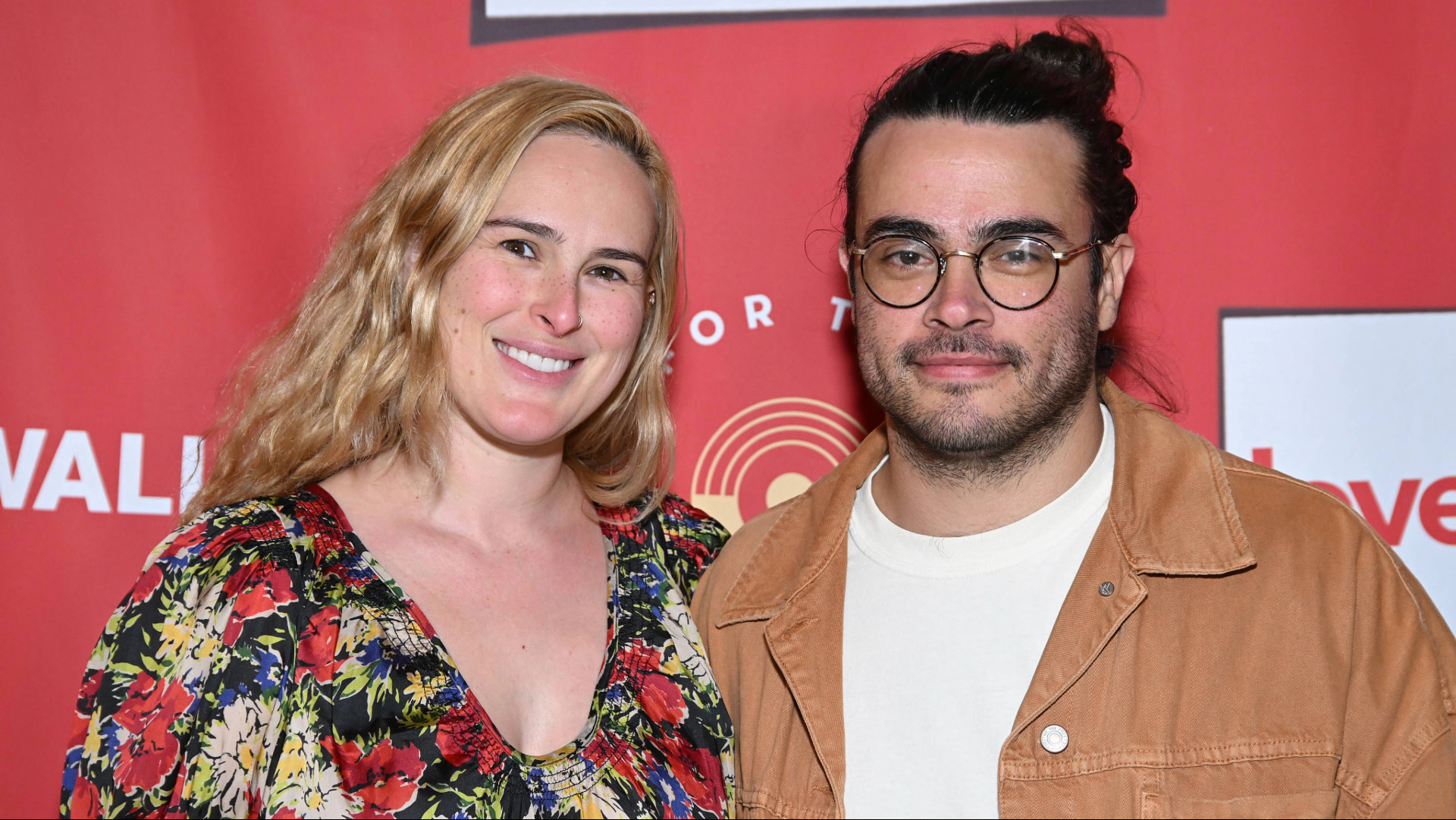 A woman in a floral dress and a man in a brown jacket pose together against a red backdrop.