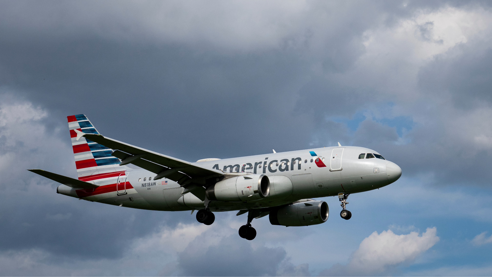 An American Airlines plane flying under a cloudy sky, with its landing gear down.