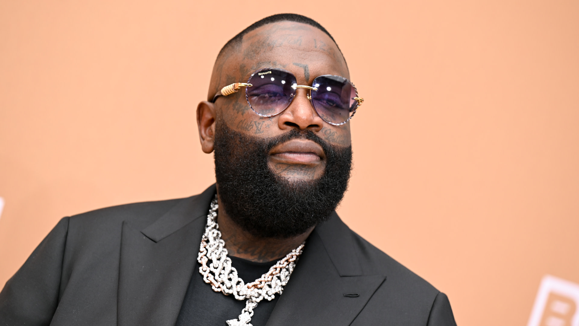 Rick Ross wearing sunglasses and a black suit, accessorized with a thick chain necklace, against a peach background.