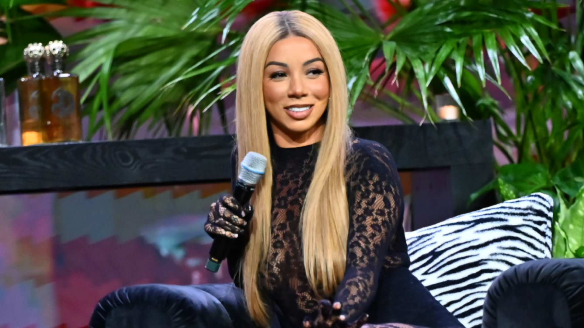 Brittany Renner with long blonde hair in a black lace outfit holds a microphone, sitting on a couch with zebra-patterned pillows.