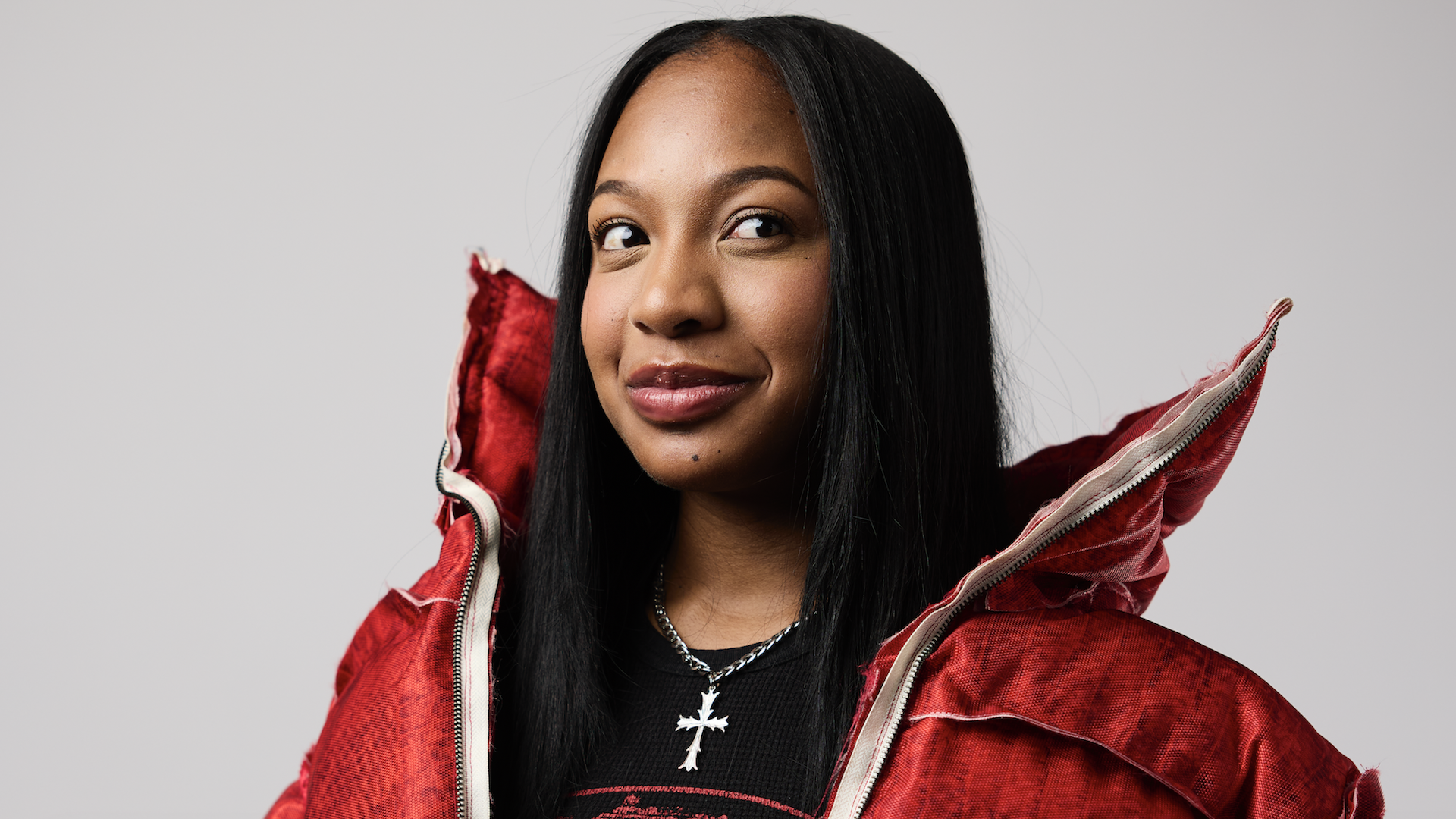 Brooklyn Frost with long black hair wearing a red jacket and a cross necklace, looking slightly to the side.