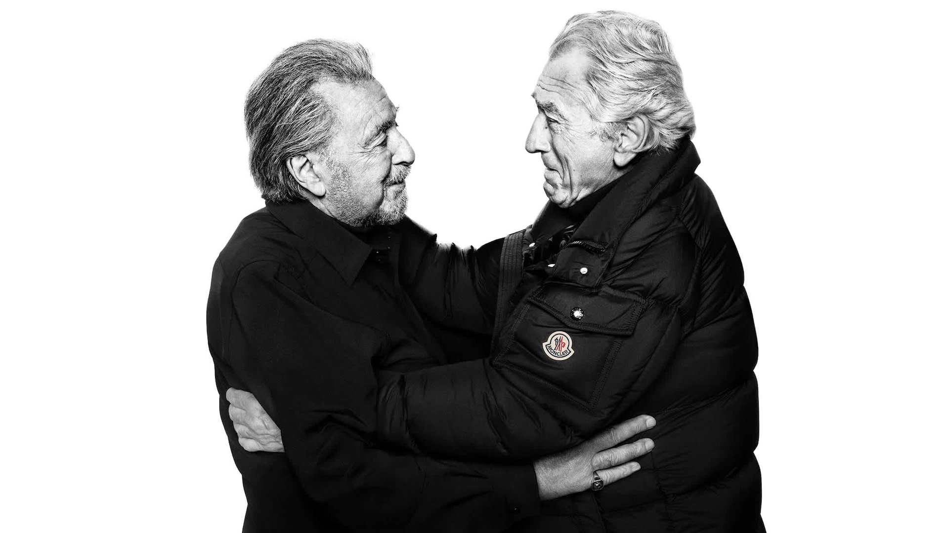 Two older men embracing in a friendly manner, both wearing dark clothing, with one in a puffy jacket. Black and white photo.