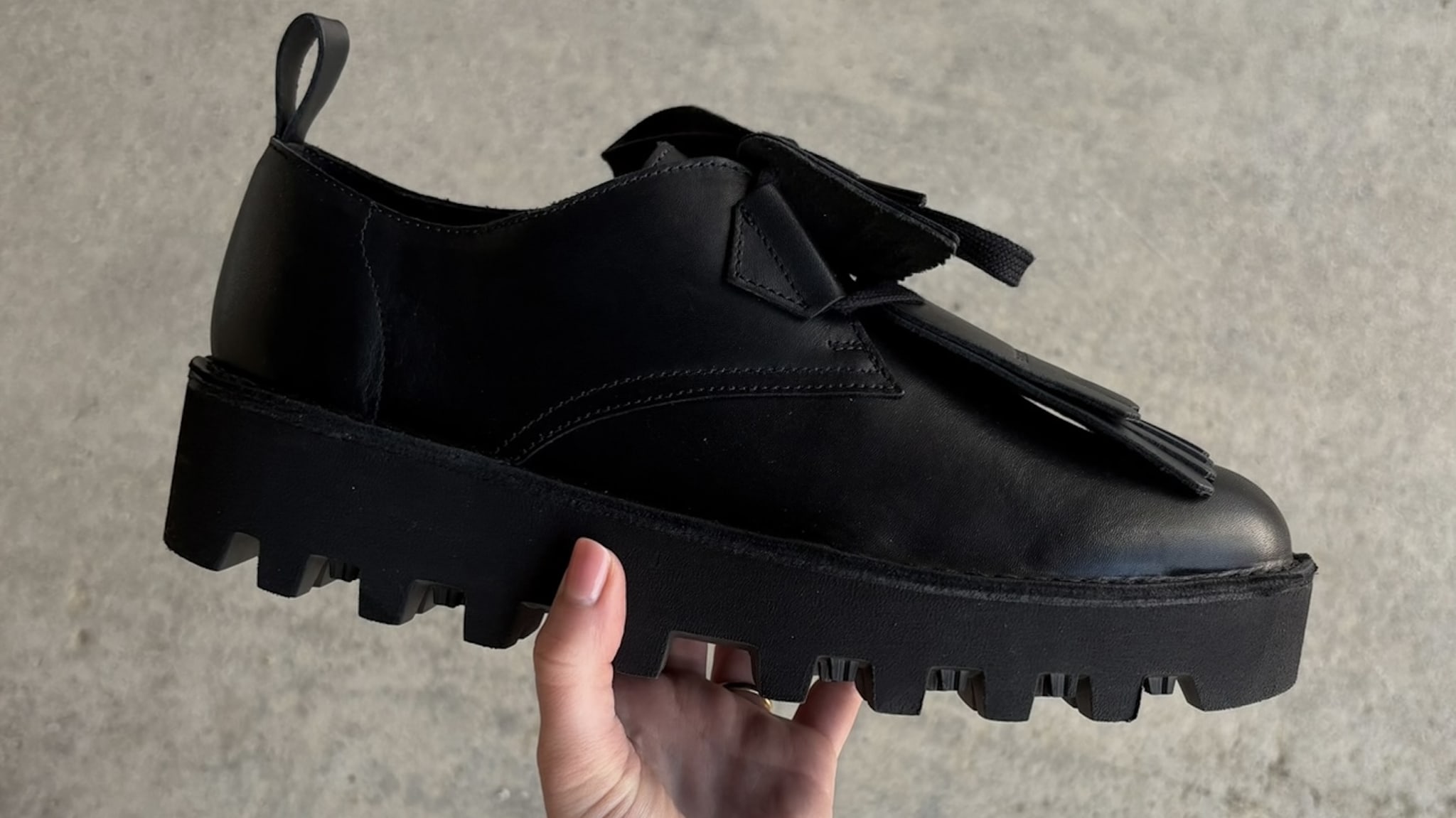 A hand holding a black leather Engineered Garments shoe with a chunky, ridged sole and decorative tassels on top.