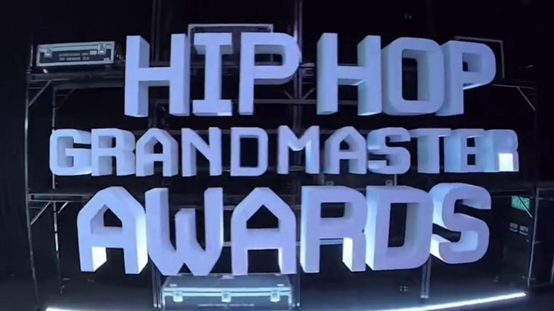 3D text reading "Hip Hop Grandmaster Awards" on a dark background with stage equipment.