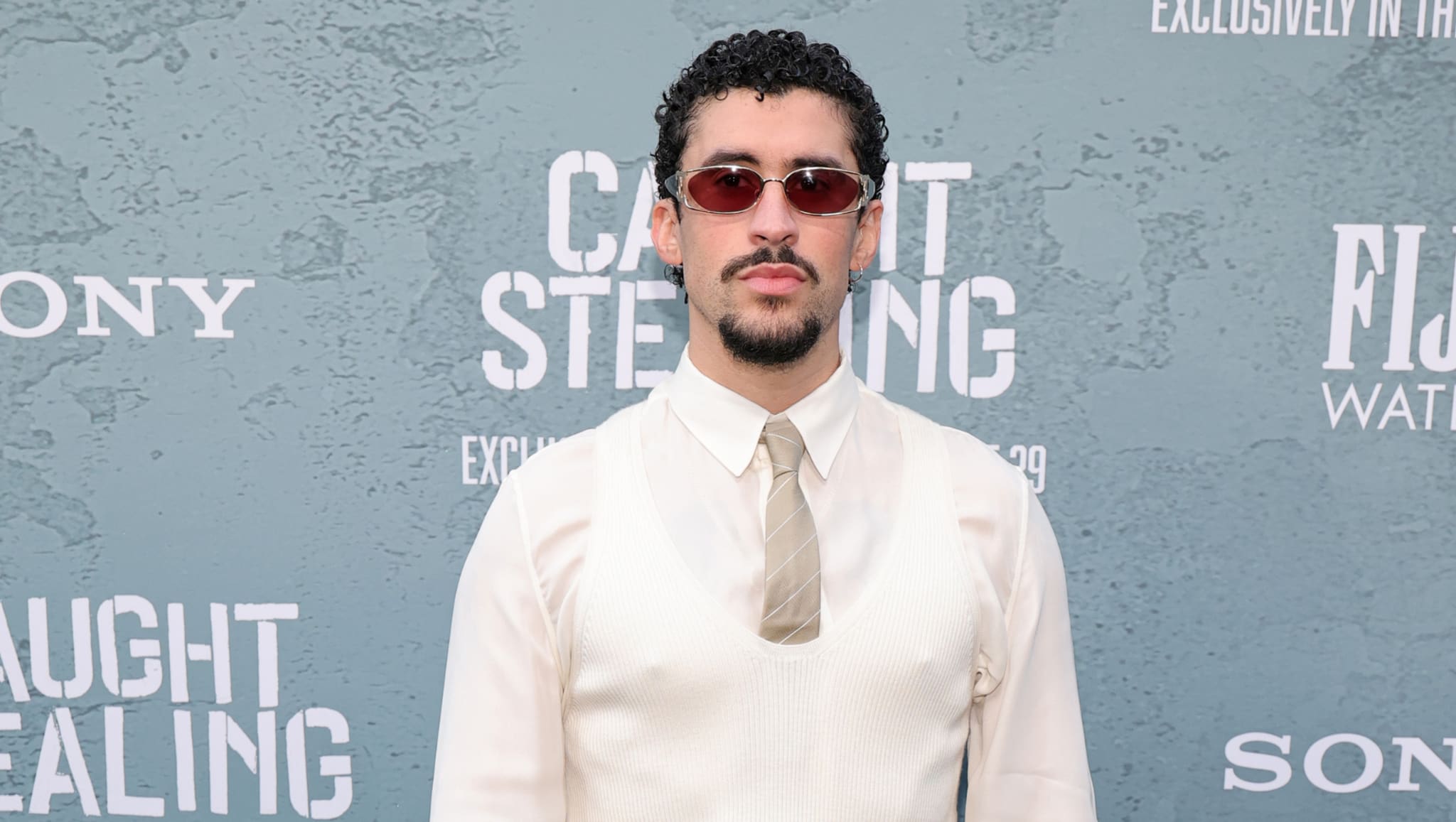 A man with curly hair and sunglasses stands in front of a backdrop with text, wearing a white shirt, tie, and vest.