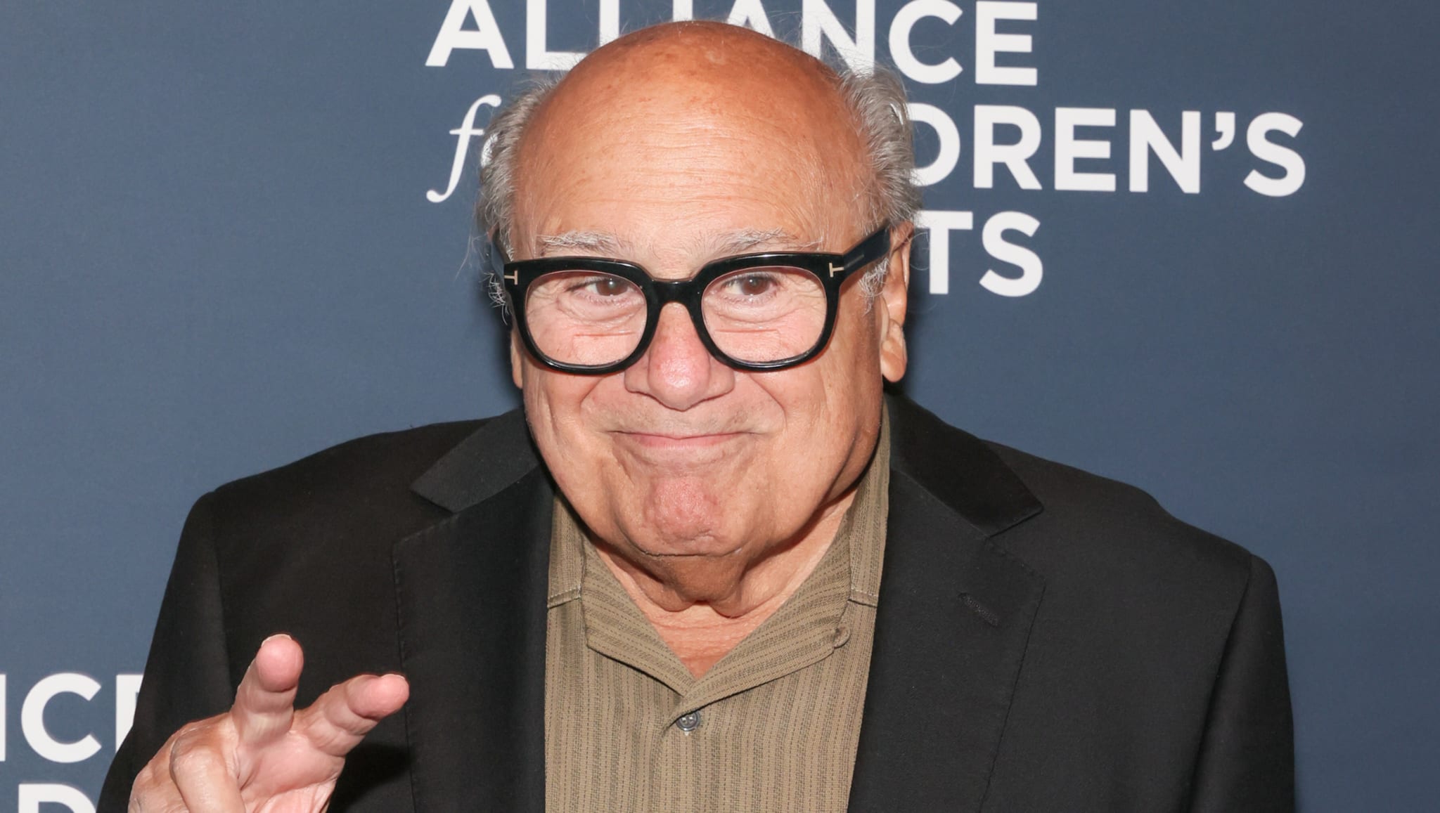 Danny DeVito, wearing glasses and a suit, poses with a peace sign at an event with a blue background.