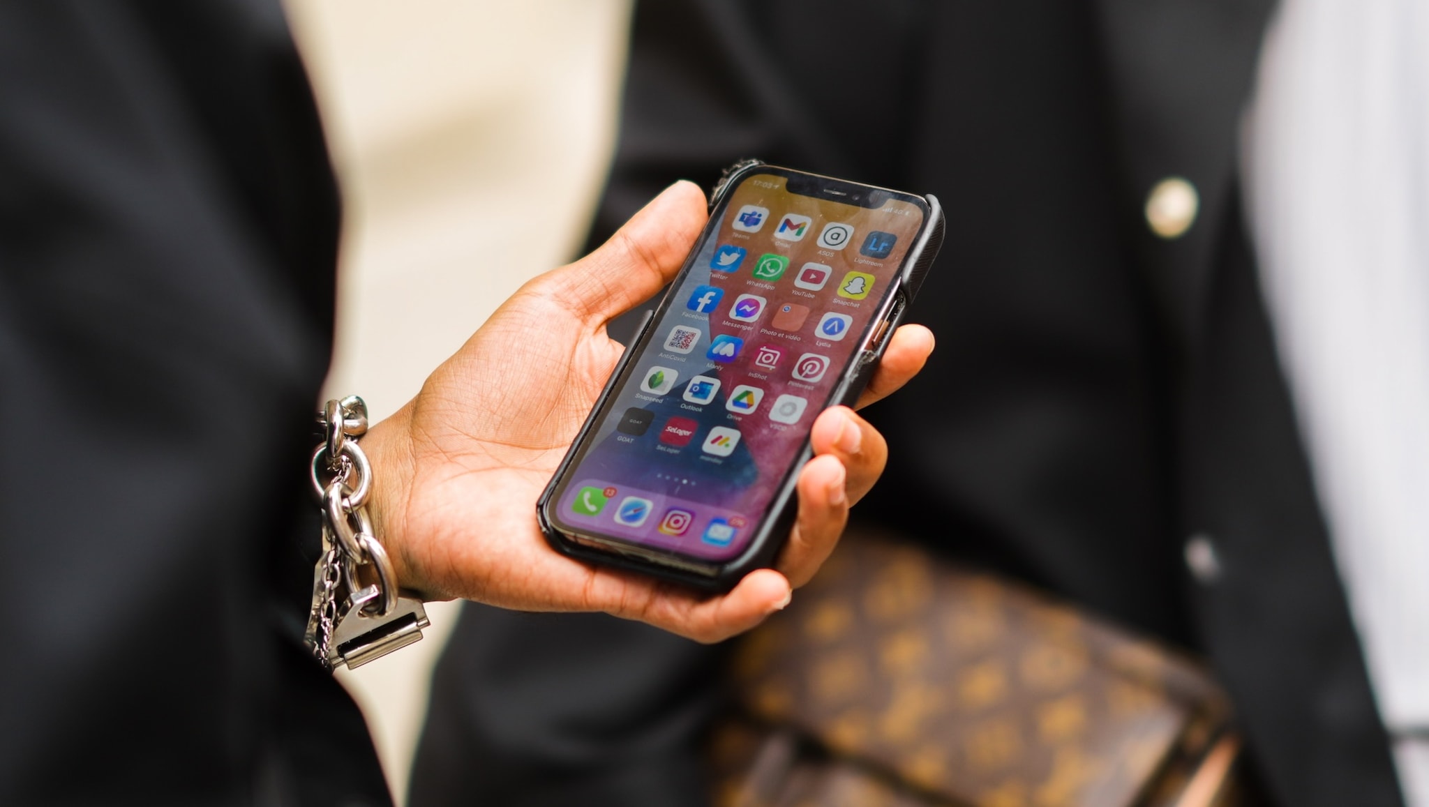 A person holding a smartphone displaying various app icons, wearing a bracelet, with a blurred background.