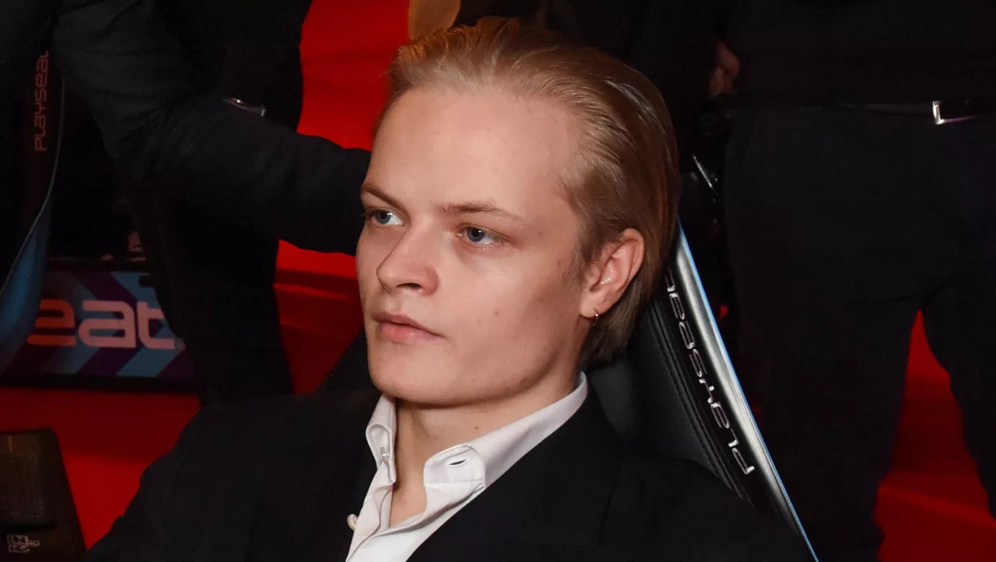 A person with blonde hair sitting on a chair, wearing a black suit and white shirt, on a red carpet background.