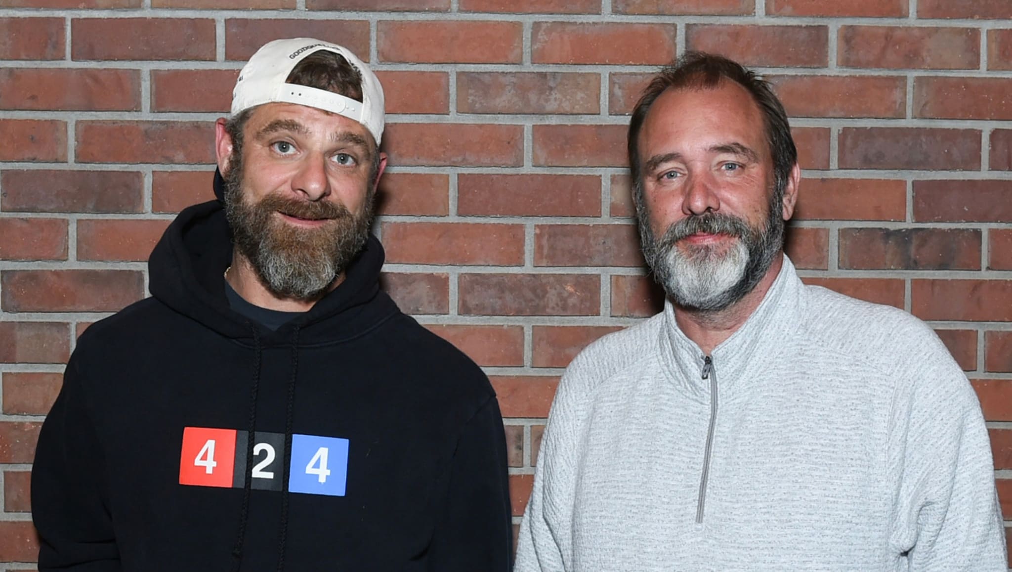 Two men with beards stand in front of a brick wall. One wears a cap and hoodie, the other a light sweater.